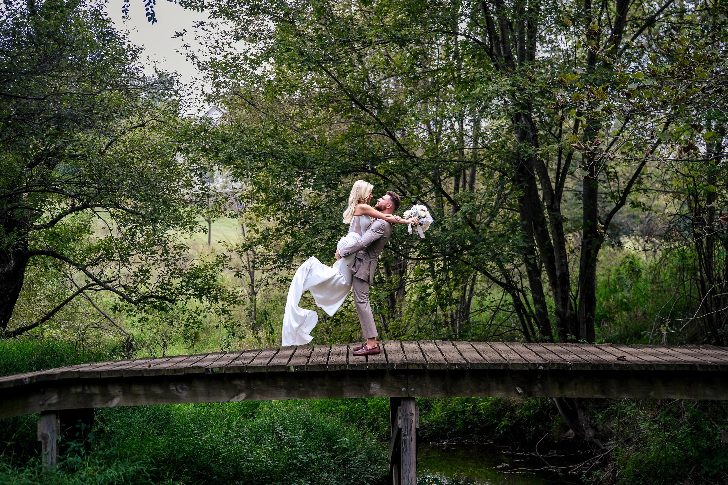 Jacob lifts Ava into an embrace on a wooden bridge surrounded by greenery.