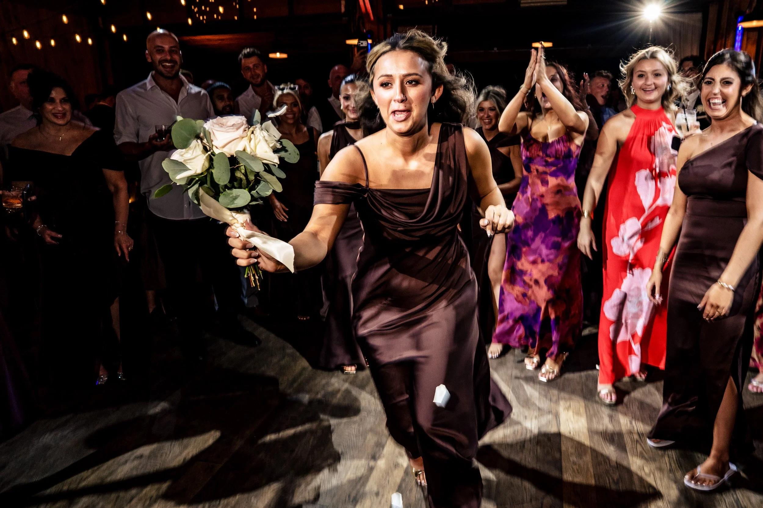 A woman in a brown dress holds the bouquet while other guests react behind her.