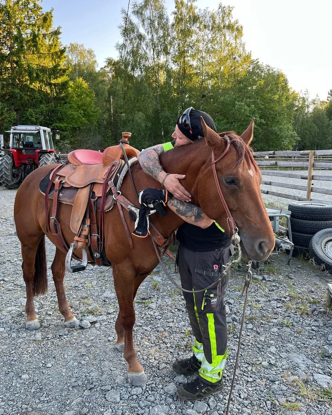 Man som kramar en häst med en sadel på, i en utomhusmiljö med träd och en traktor i bakgrunden.