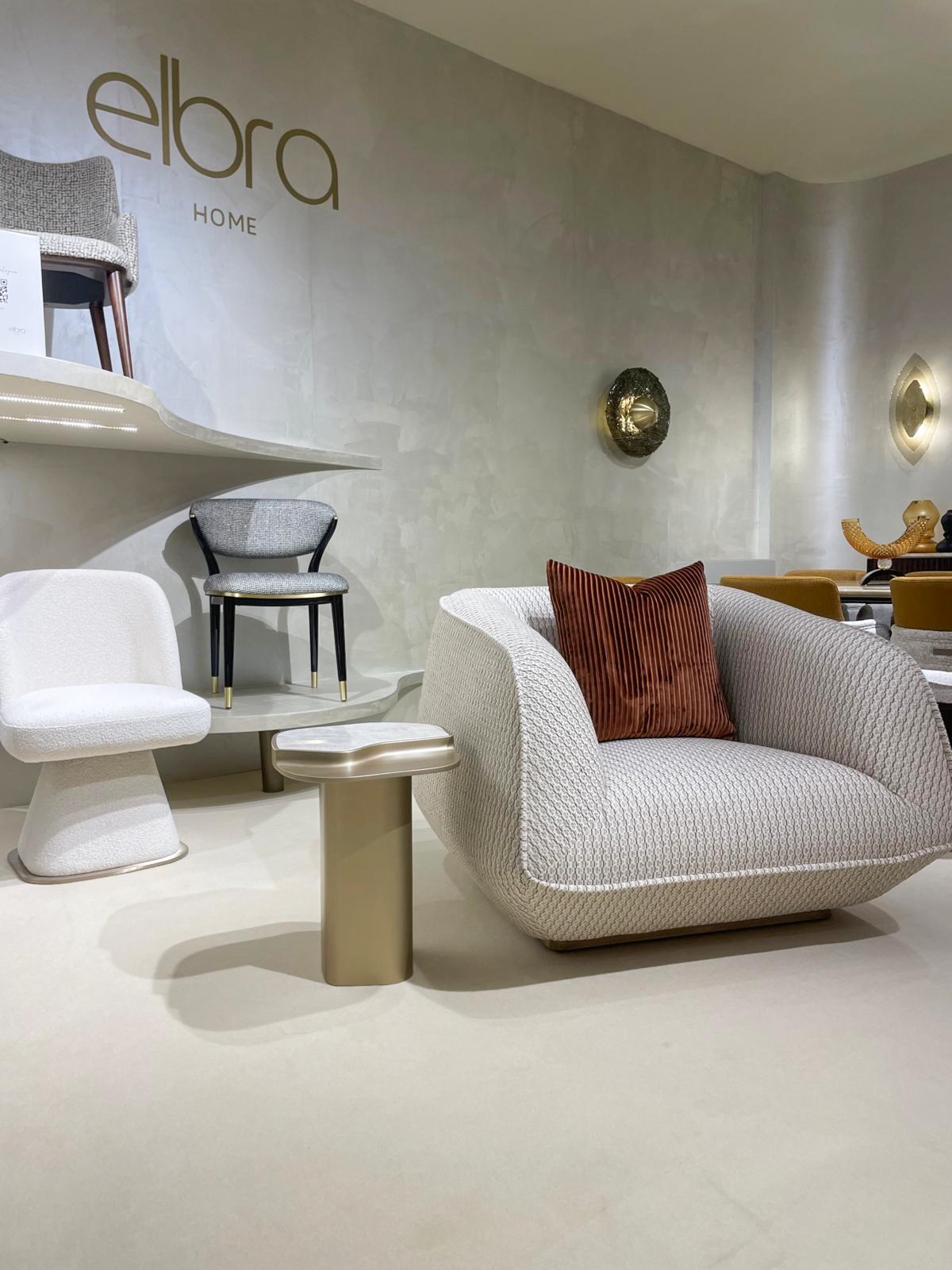 Luxury furniture exhibition stand at Salone del Mobile featuring a mid-century modern dining room, a fluted wood private bar with marble accents, and a sculptural chair gallery in a warm, textured interior.