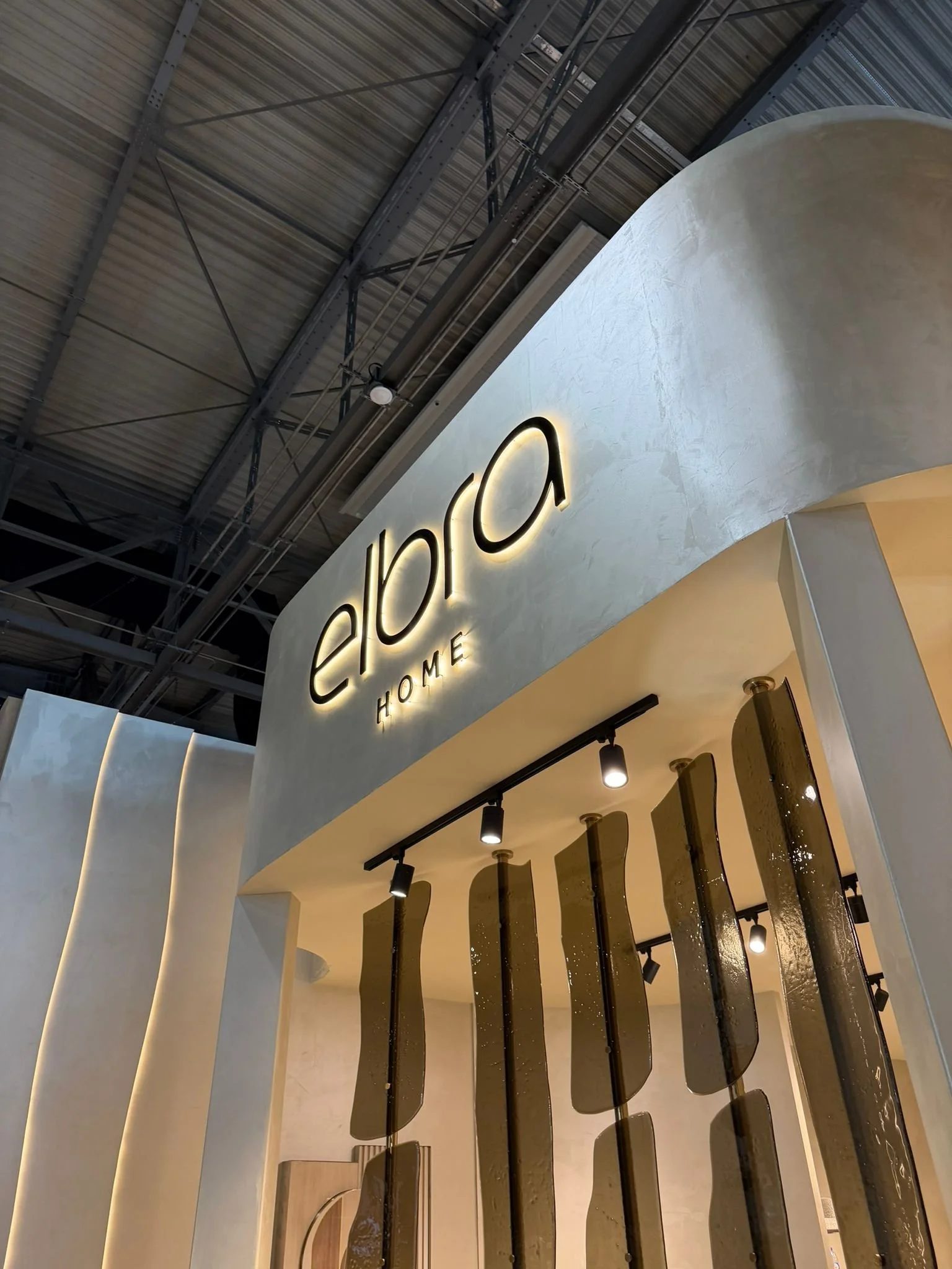 Luxury furniture exhibition stand at Salone del Mobile featuring a mid-century modern dining room, a fluted wood private bar with marble accents, and a sculptural chair gallery in a warm, textured interior.