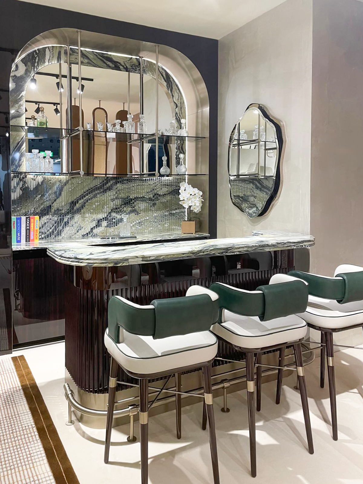 Luxury furniture exhibition stand at Salone del Mobile featuring a mid-century modern dining room, a fluted wood private bar with marble accents, and a sculptural chair gallery in a warm, textured interior.