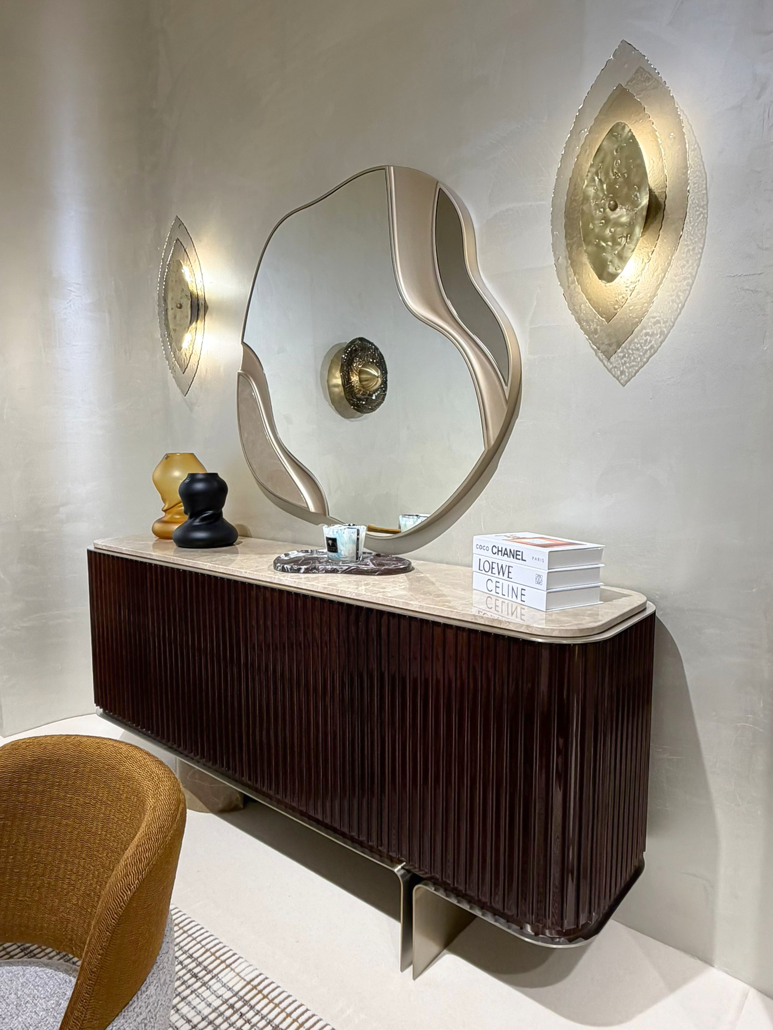 Luxury furniture exhibition stand at Salone del Mobile featuring a mid-century modern dining room, a fluted wood private bar with marble accents, and a sculptural chair gallery in a warm, textured interior.