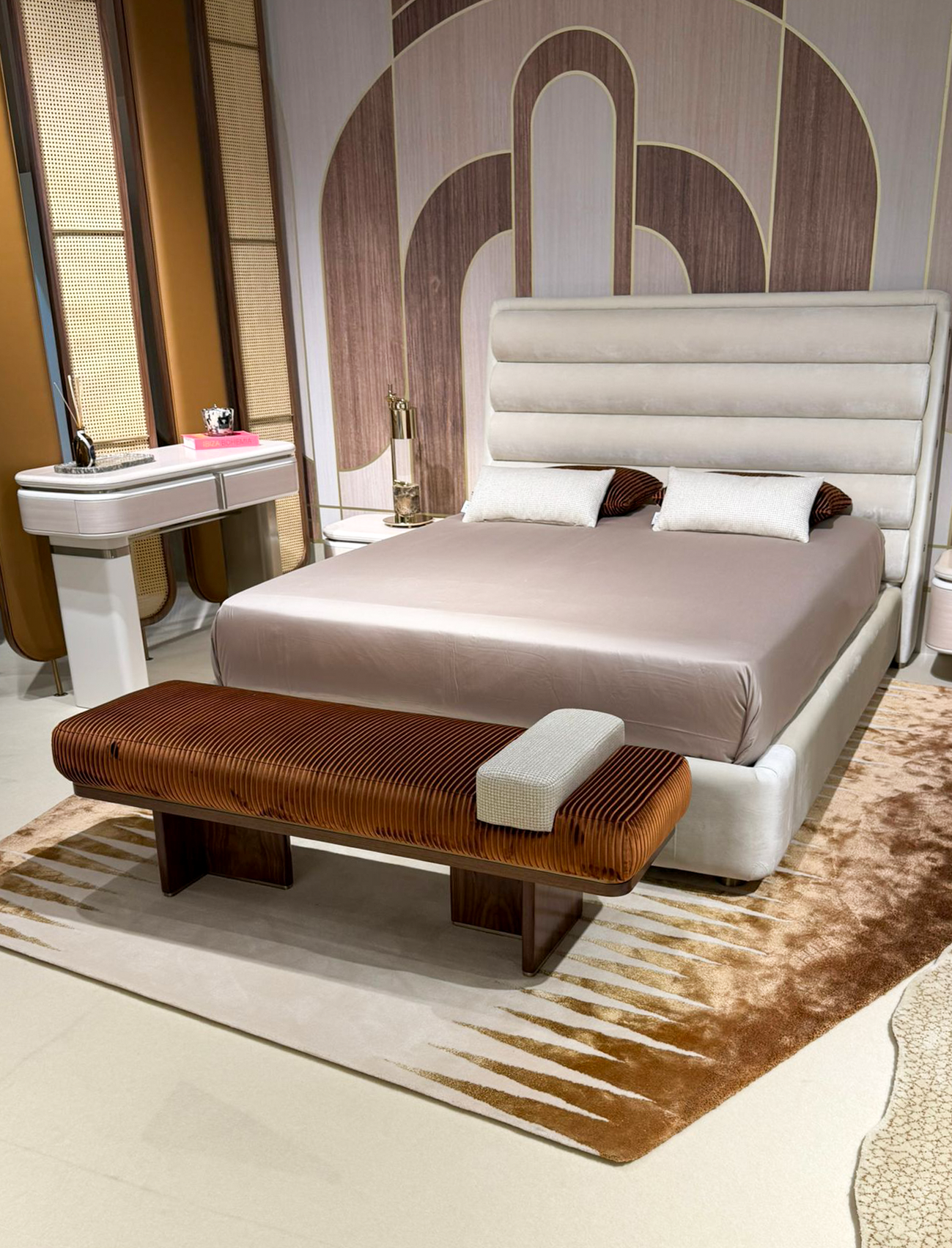 Luxury furniture exhibition stand at Salone del Mobile featuring a mid-century modern dining room, a fluted wood private bar with marble accents, and a sculptural chair gallery in a warm, textured interior.