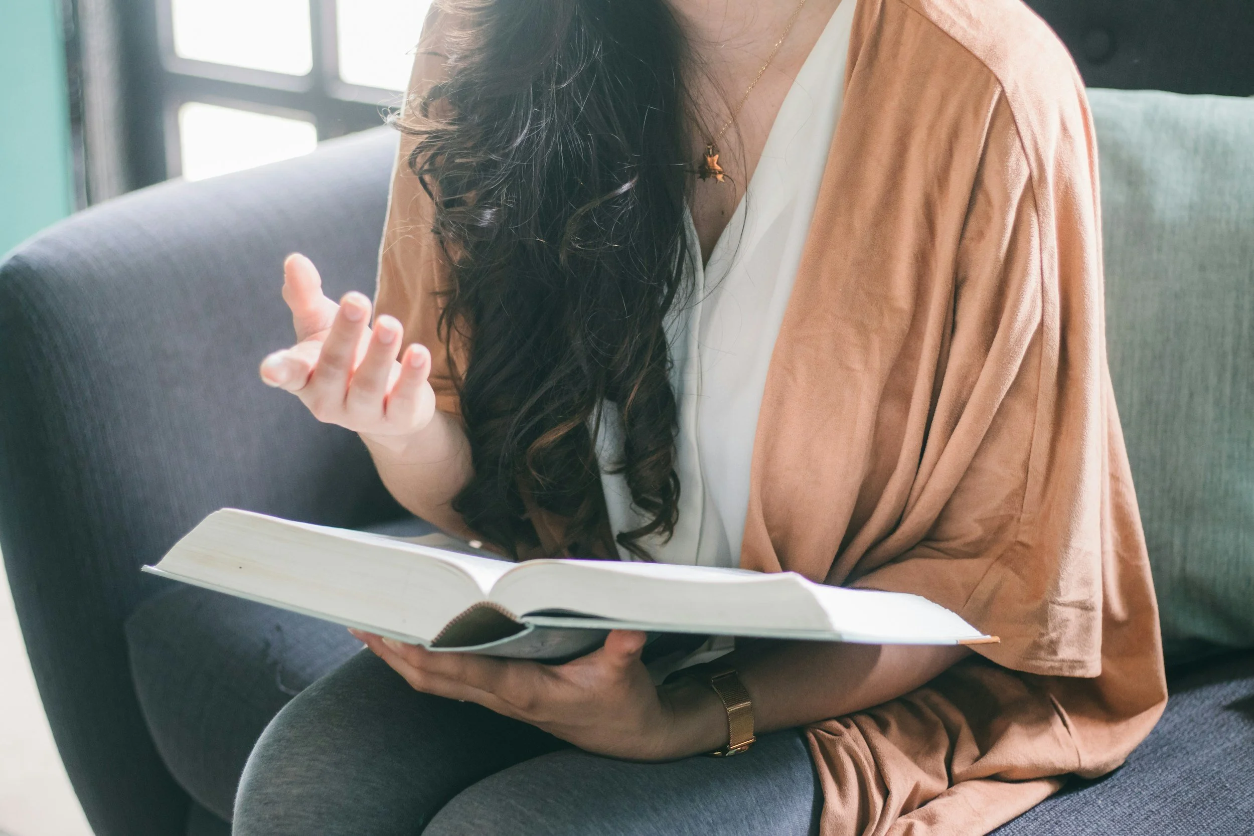 A woman with long dark curly hair reading a book while sitting on a gray chair in a well-lit room.