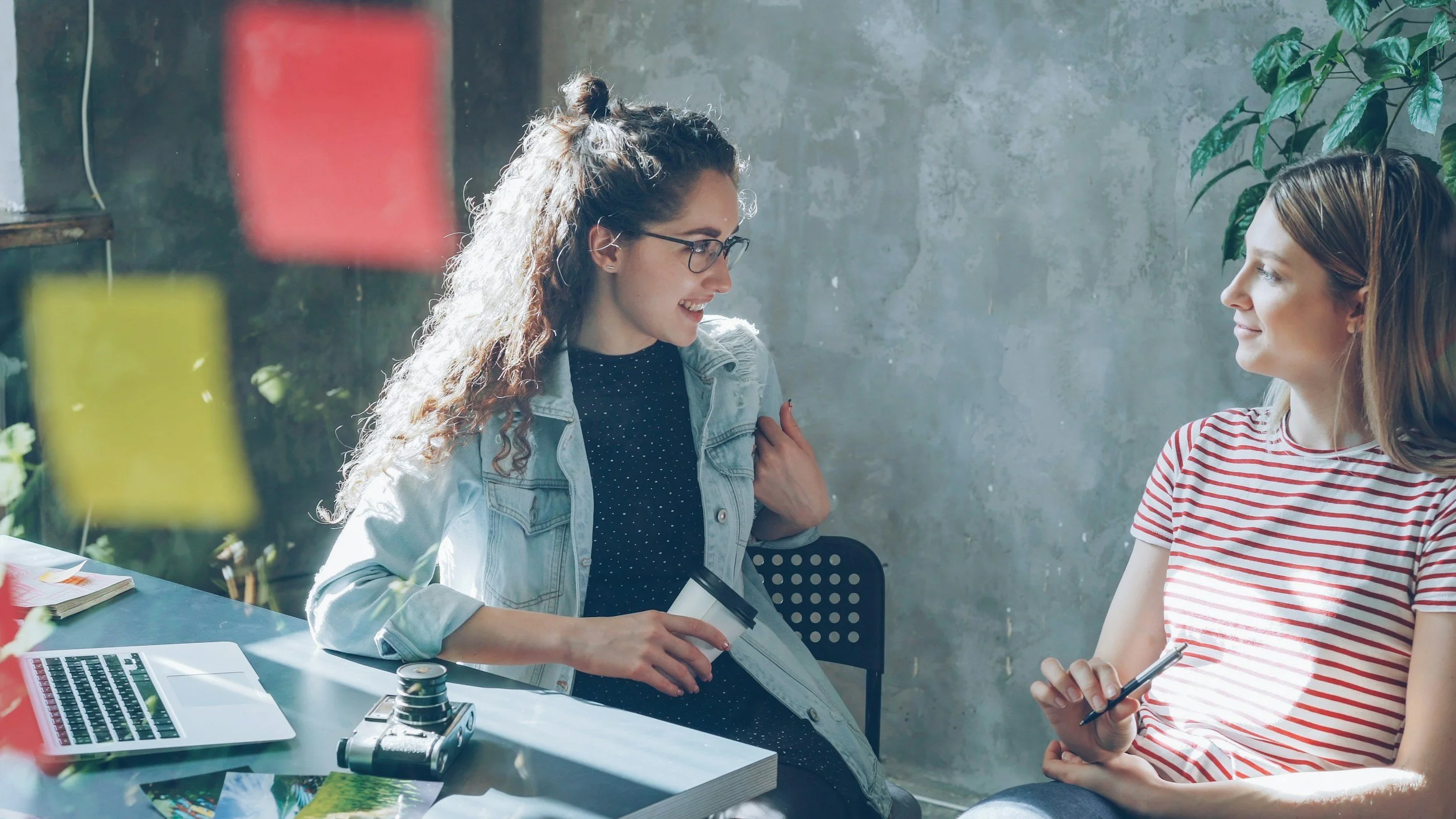 Two women sitting at a table and engaging in conversation. One woman has curly hair, glasses, and a denim jacket, holding a paper cup. The other woman has straight hair and is holding a smartphone. The table has a laptop, a notebook, and a camera. Sunlight streams through a window, illuminating the scene.
