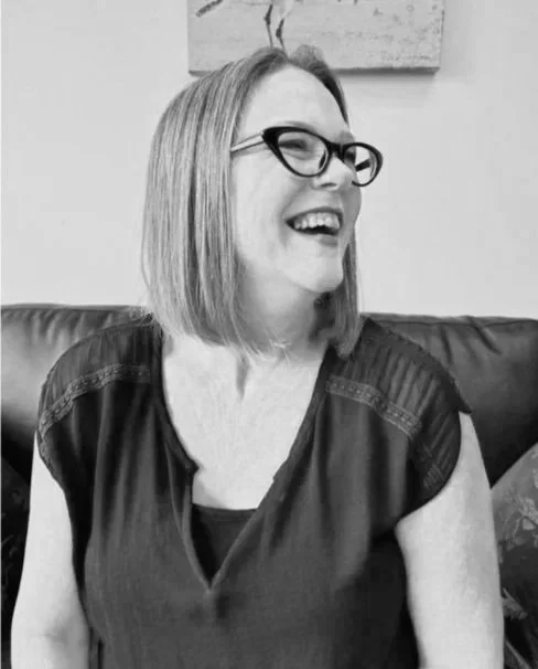 A woman with shoulder-length hair, wearing glasses and a dark blouse, smiling and sitting on a sofa in a room with wall art in the background.