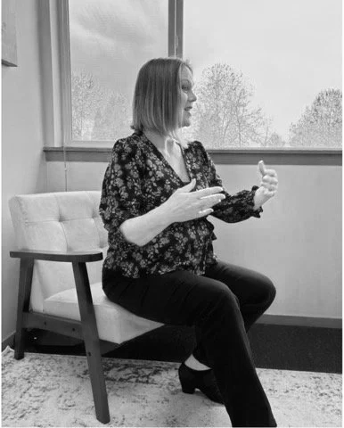 A woman sitting on an office chair in front of a window, gesturing with her hands.