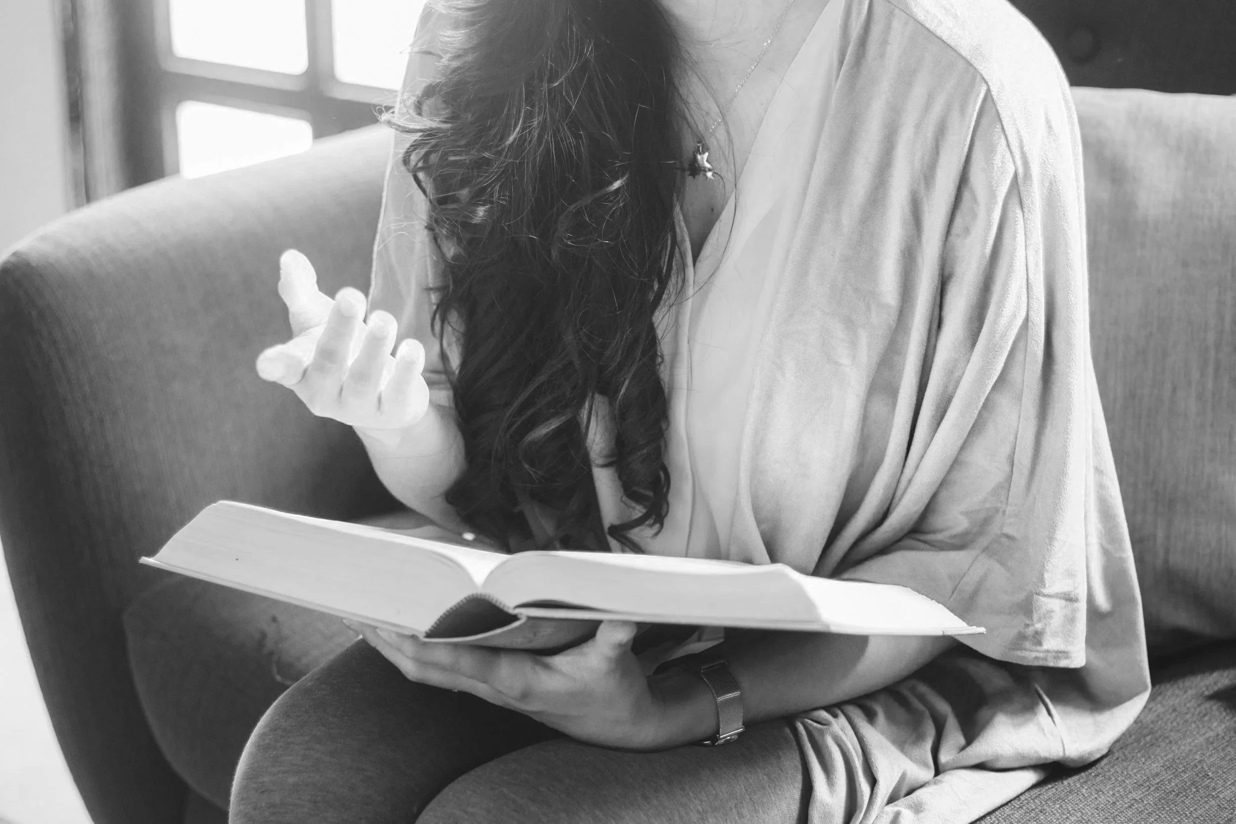 A woman sitting on a couch reading a book, with long curly hair, wearing a loose shirt and a watch, in a black and white photo.