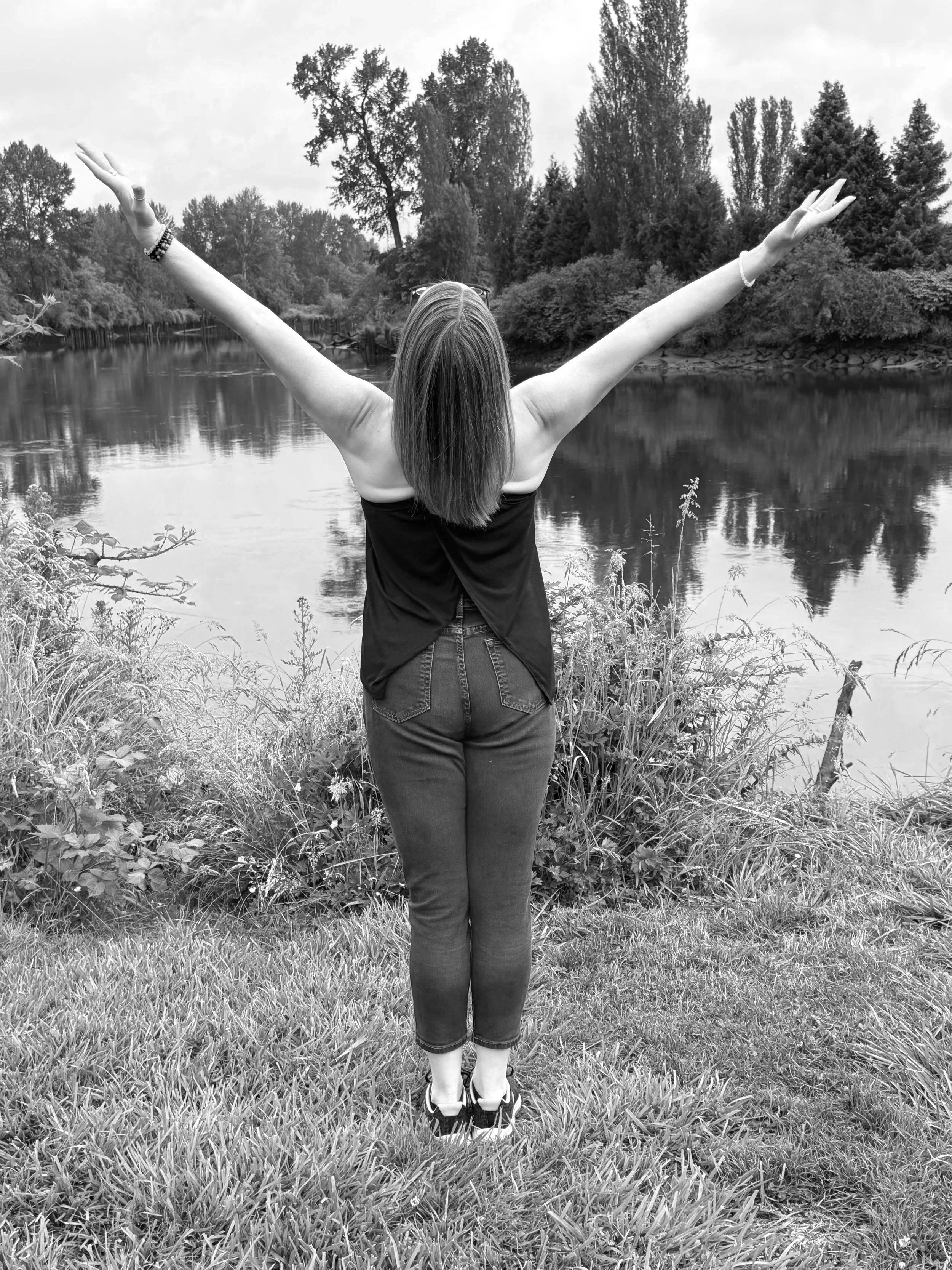 A woman stands on grass by a river with her arms raised wide, facing away from the camera, with trees in the background.