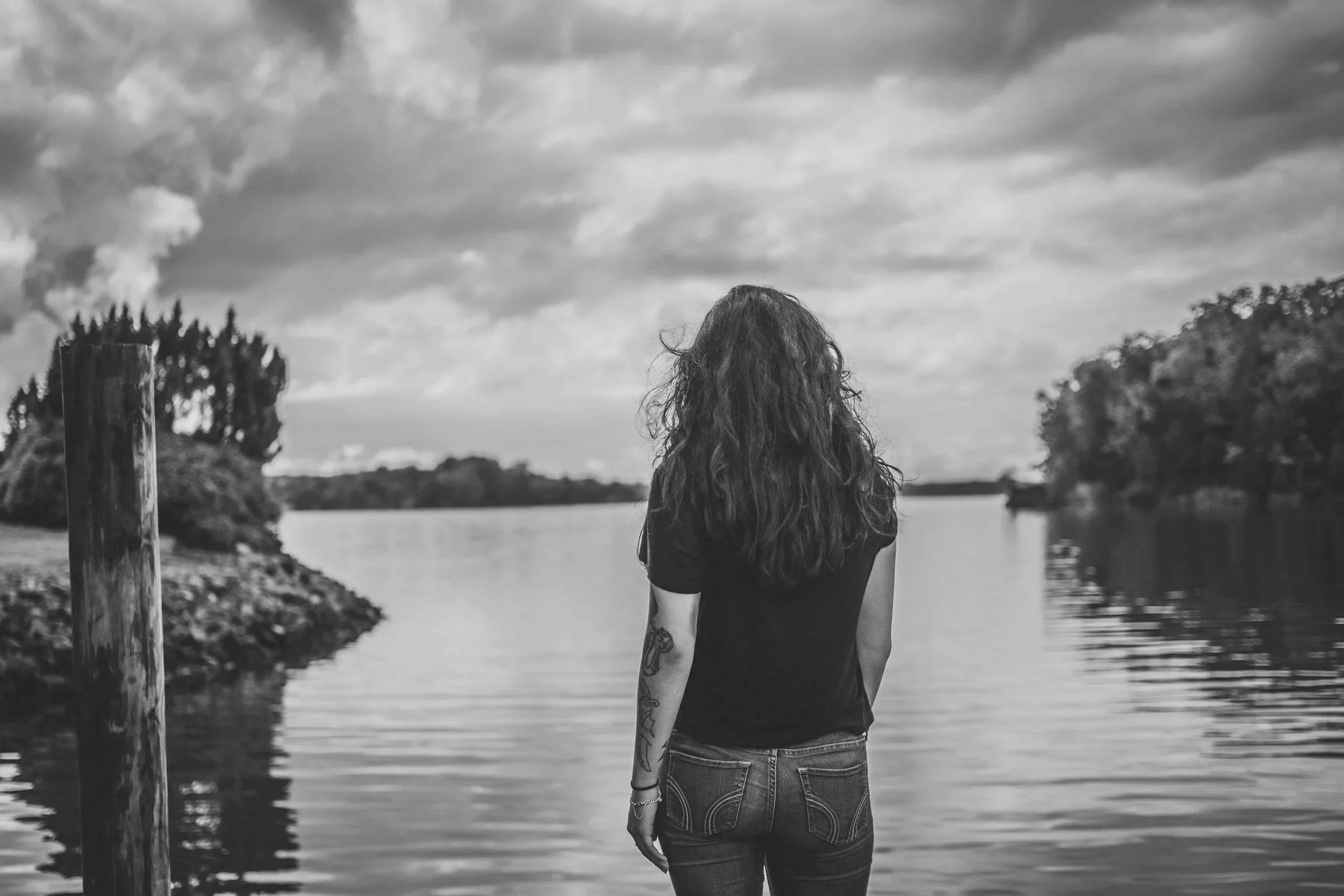 A woman with long curly hair stands by a river, facing away, with trees and cloudy sky in the background, in a black and white photo.