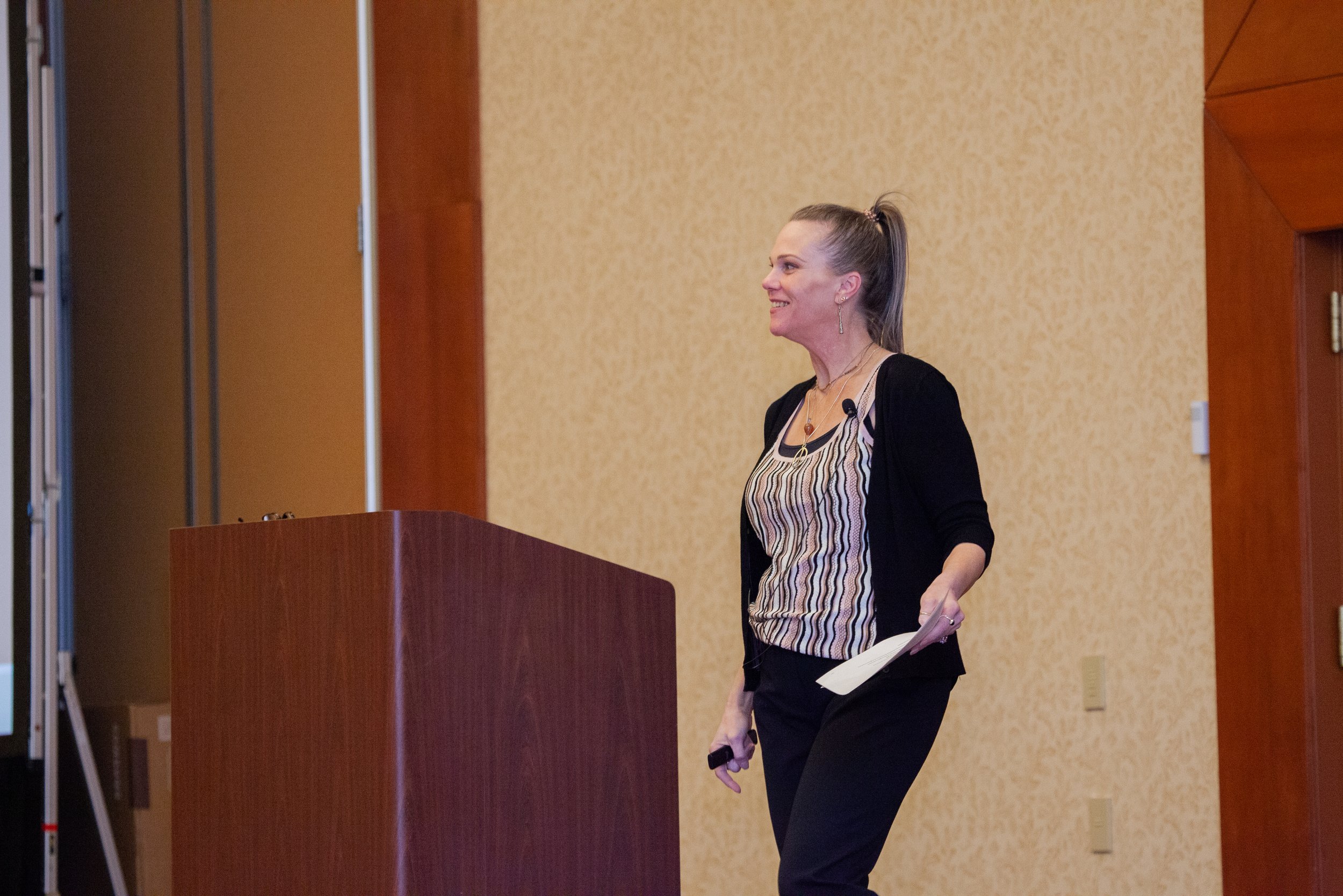 A woman with blonde hair tied in a high ponytail, wearing a black cardigan over a striped blouse, stands next to a podium in a conference room. She is smiling and holding papers and a remote control in her hands.