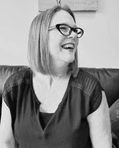A woman with shoulder-length hair, wearing glasses and a dark top, smiling and sitting on a couch.