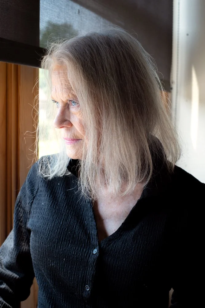 A middle-aged woman with long gray hair and blue eyes looking out a window, wearing a black textured button-up shirt, standing indoors near a wooden window frame.