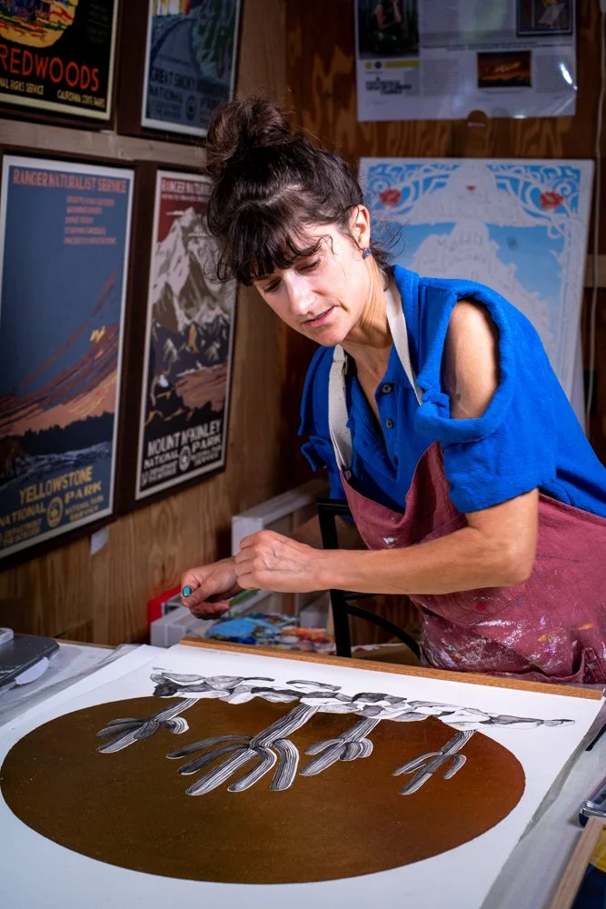 A woman with dark hair in a bun wearing a blue shirt and pink apron working on an art project at a desk in a room decorated with vintage travel posters.