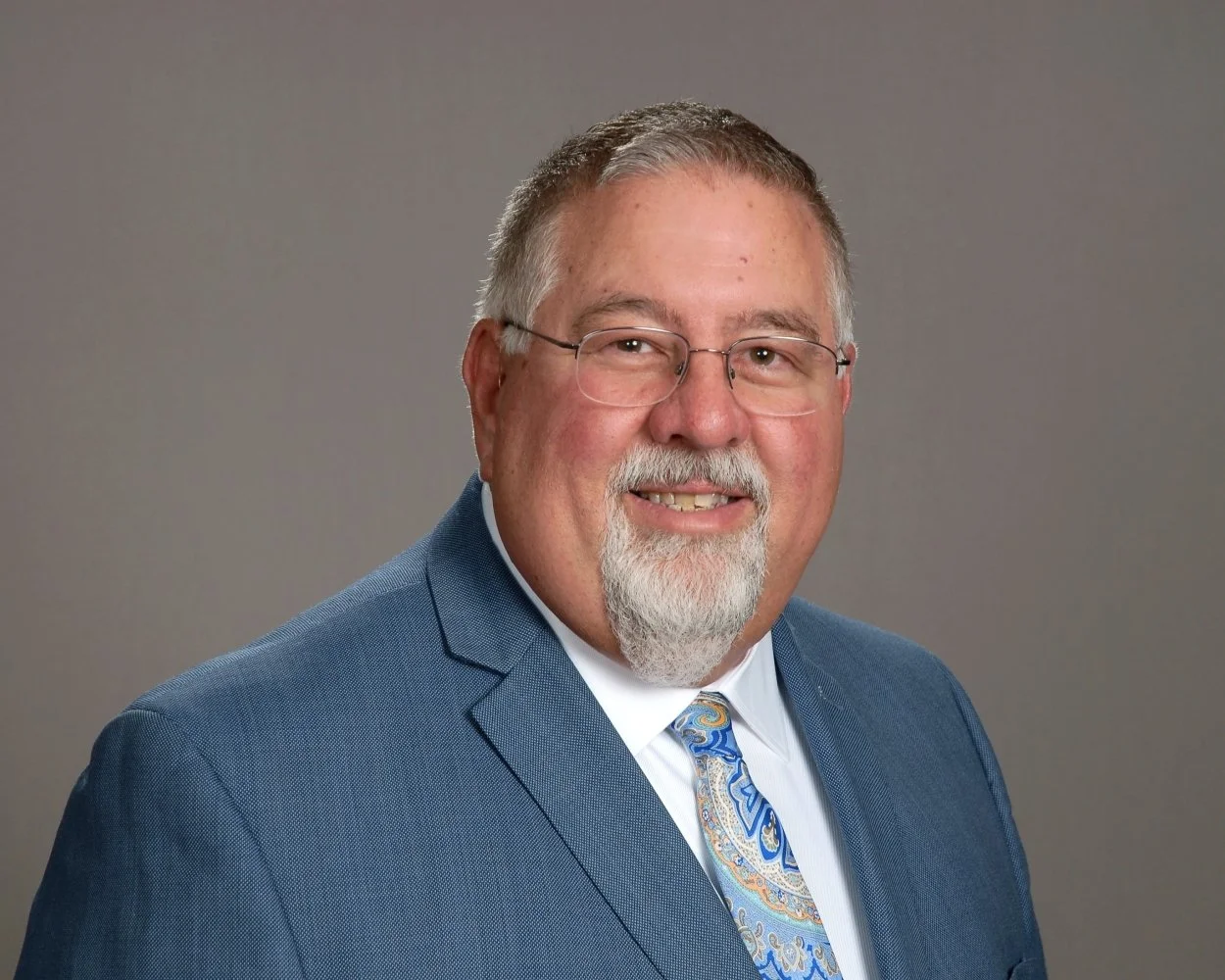 A professional man with glasses, a beard, and white hair, wearing a dark suit, light shirt, and blue tie