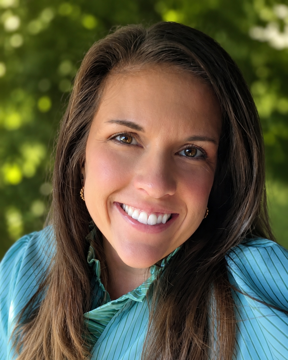 A woman with dark hair and a bright smile, wearing a blue top and a gold necklace.