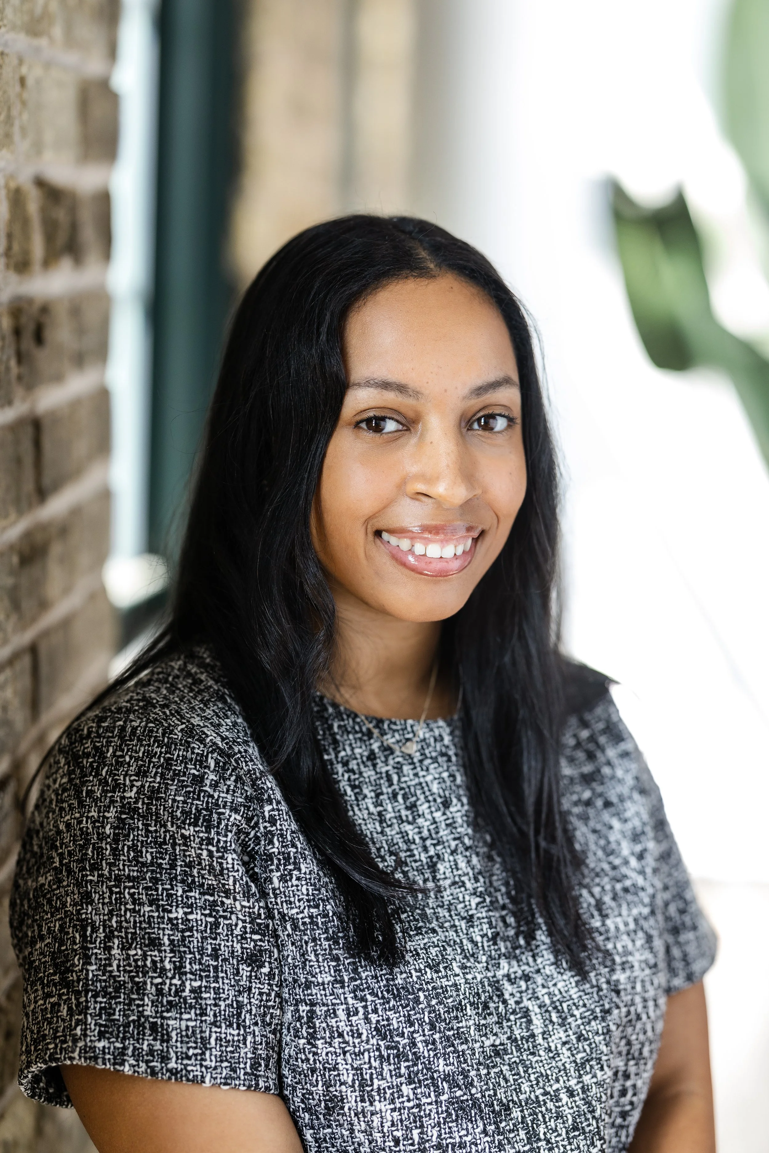 Young woman with long black hair smiling indoors near a brick wall and a large plant.