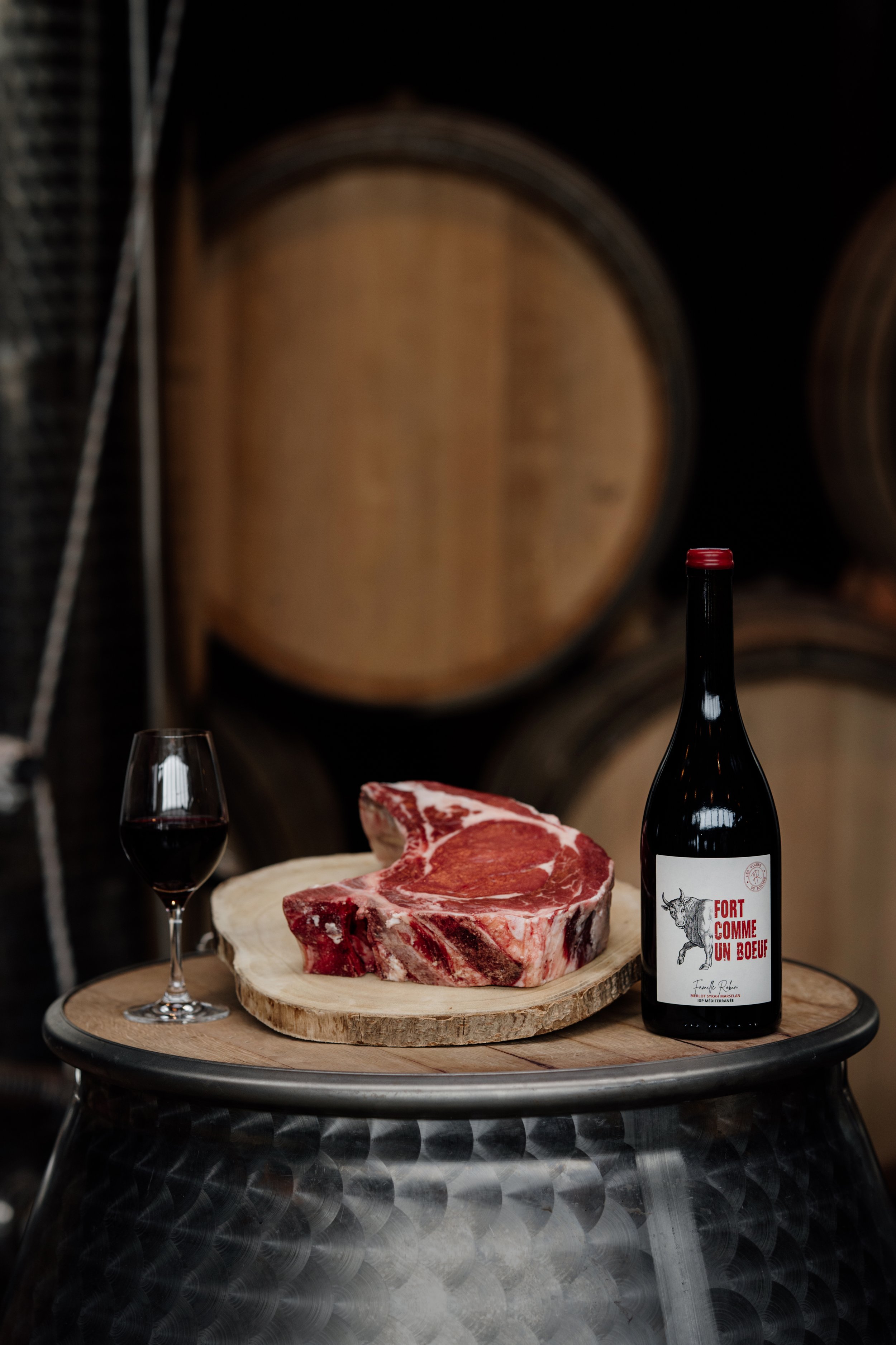 Un morceau de viande crue sur une planche en bois avec une bouteille de vin rouge intitulée "Fort Comme Un Bœuf" et un verre de vin rouge, posés sur un tonneau.