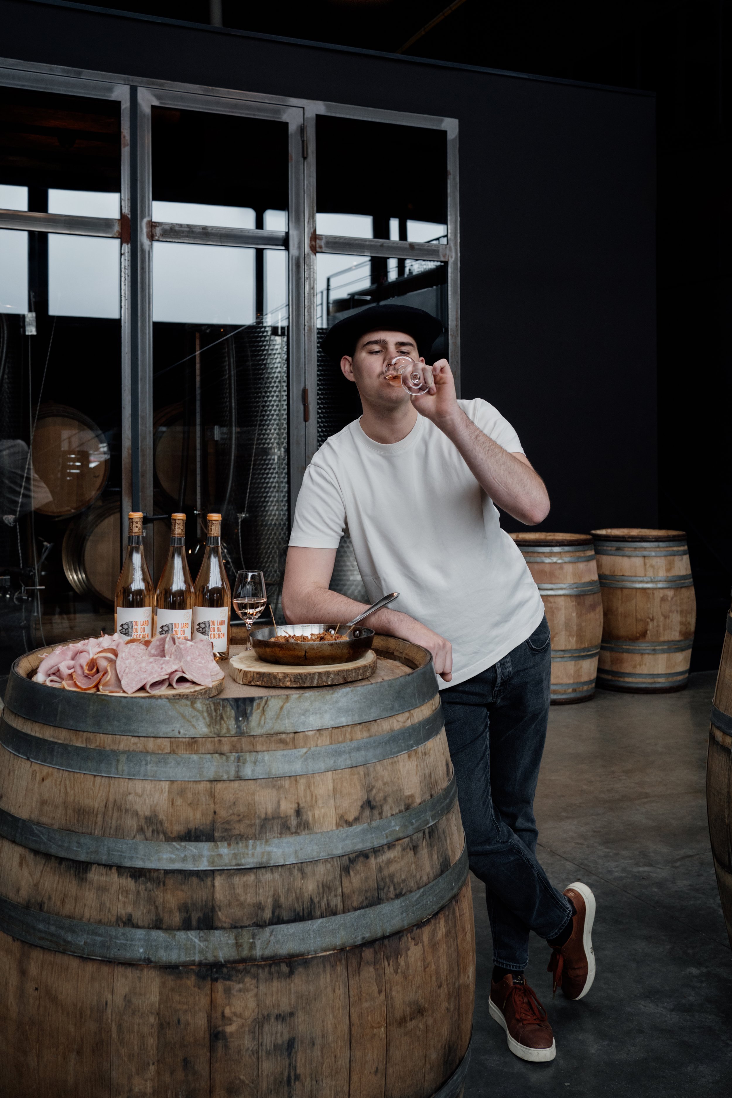 Un homme portant un chapeau noir et un t-shirt blanc déguste un verre de vin ou de boisson alcoolisée dans une cave à vin, s'appuyant sur un tonneau en bois. Sur le tonneau, il y a des plats de charcuterie et des bouteilles de vin. En arrière-plan, o