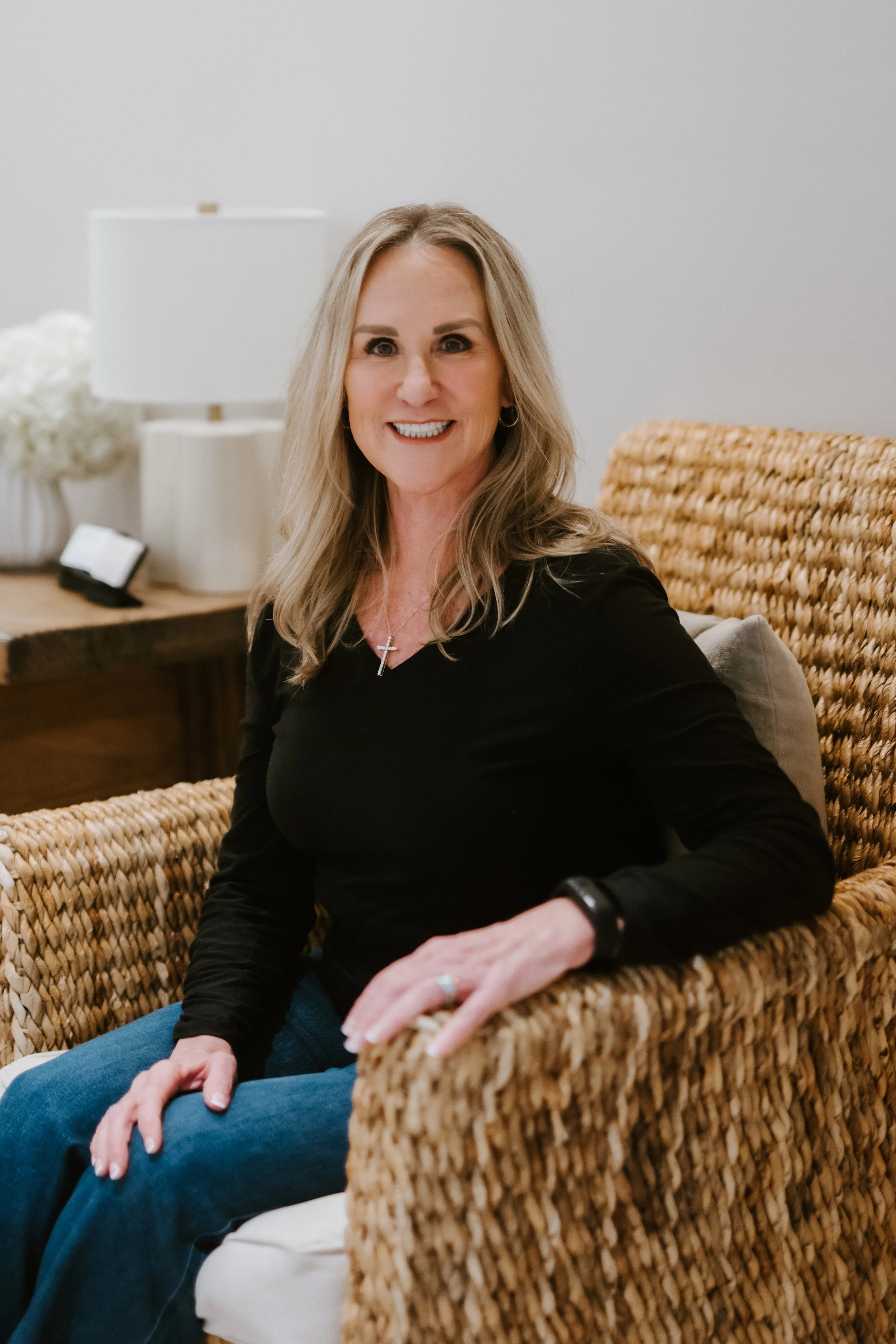 A woman with blonde hair, wearing a black top, sitting on a woven chair, smiling at the camera in a cozy indoor setting.