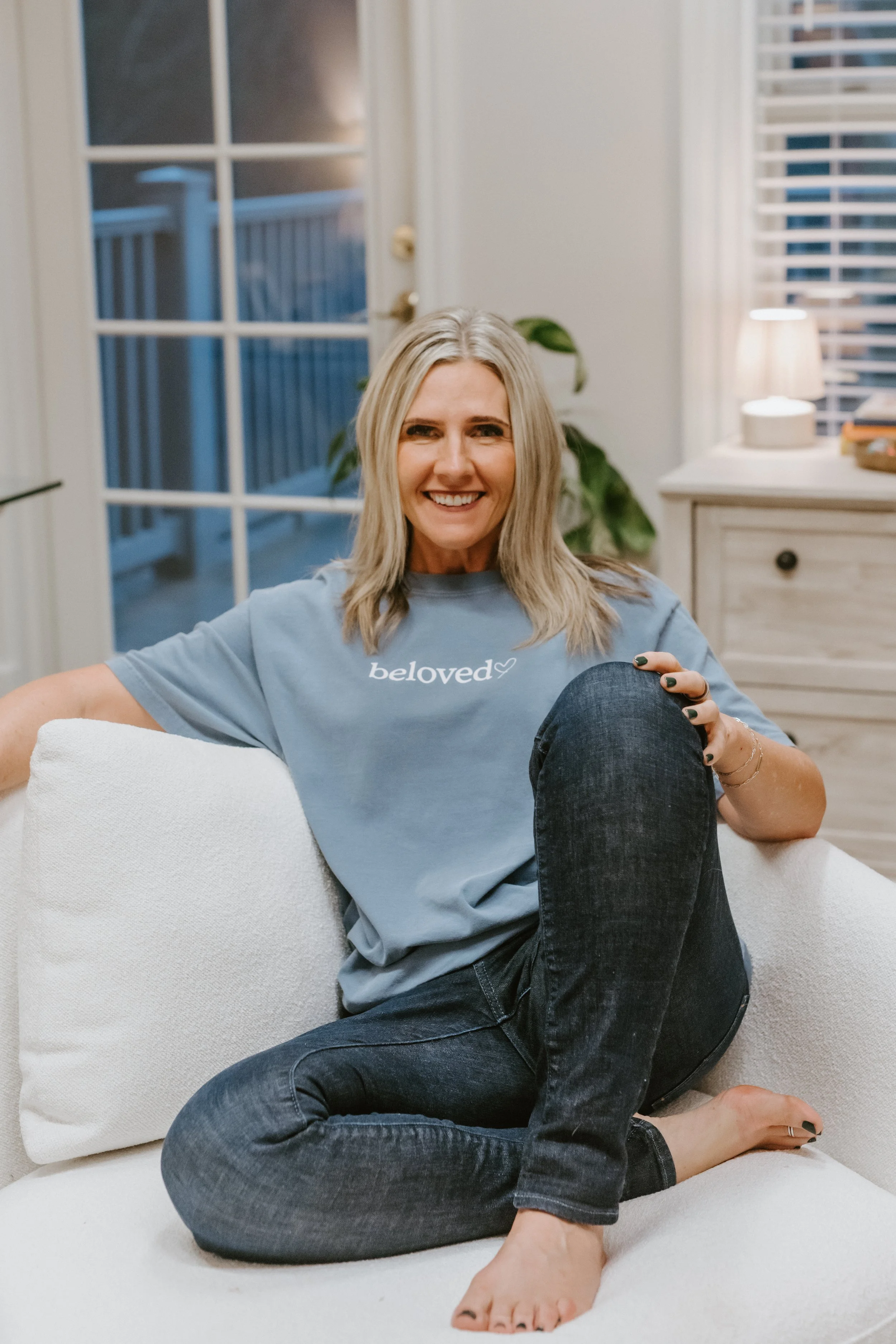 A smiling woman with gray hair sitting on a white couch in a cozy, well-lit room.