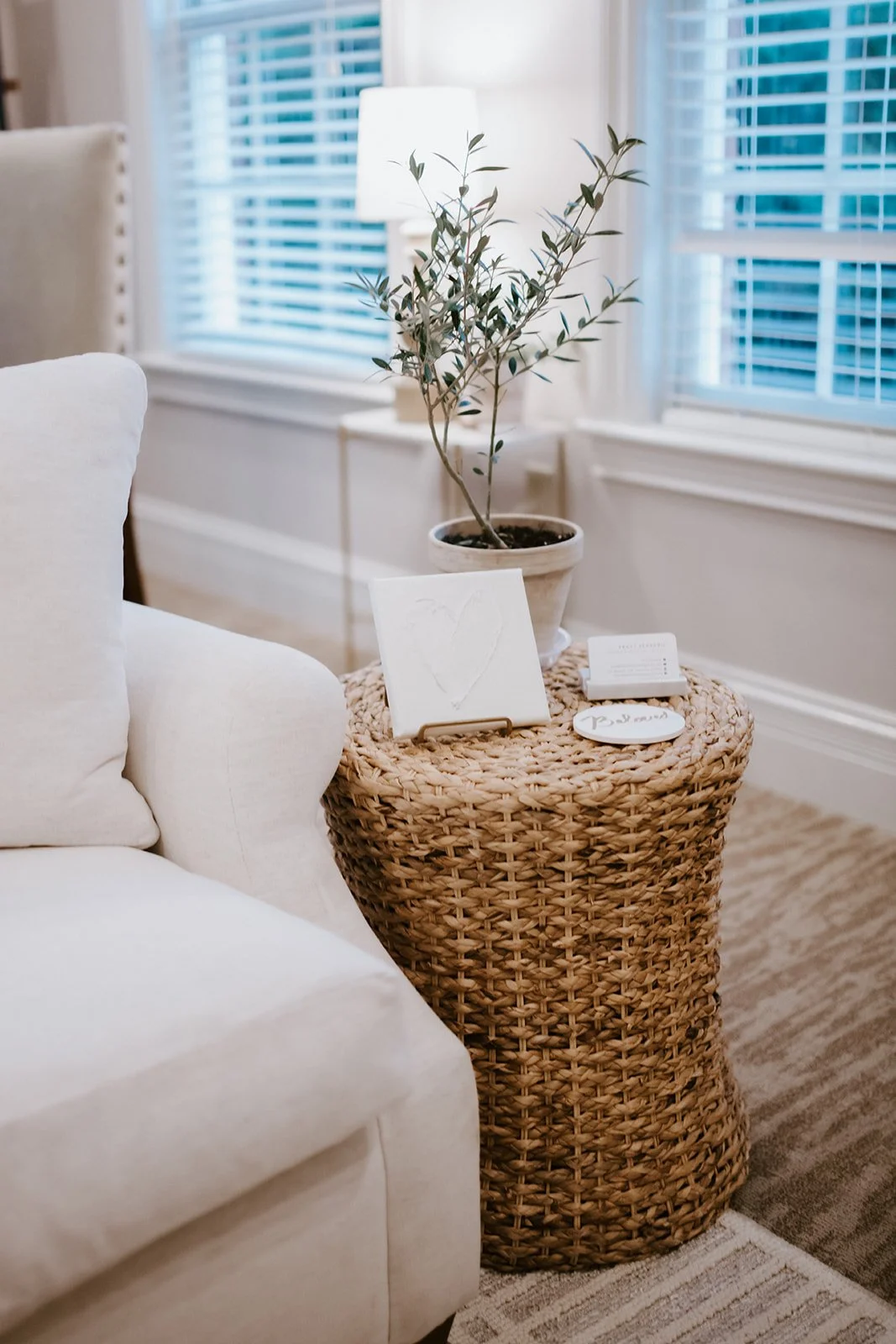 A woven basket side table holding a potted plant, a framed heart-shaped art piece, and small decorative items, next to a white sofa with beige pillows in a well-lit living room.