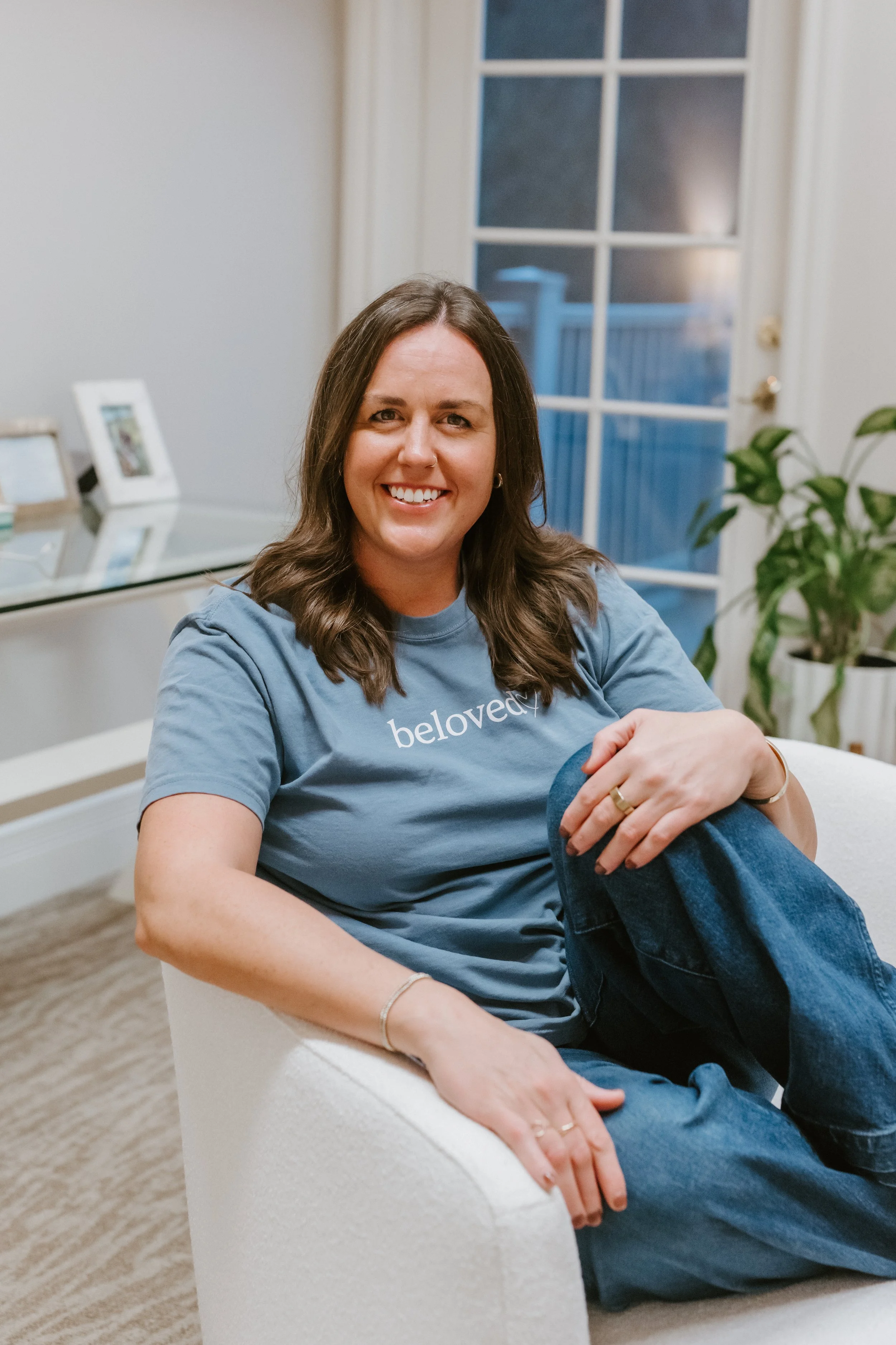 A woman with long brown hair, smiling, sitting on a white armchair in a room with a glass door and potted plants, wearing a gray t-shirt with the word 'beloved' on it and jeans.
