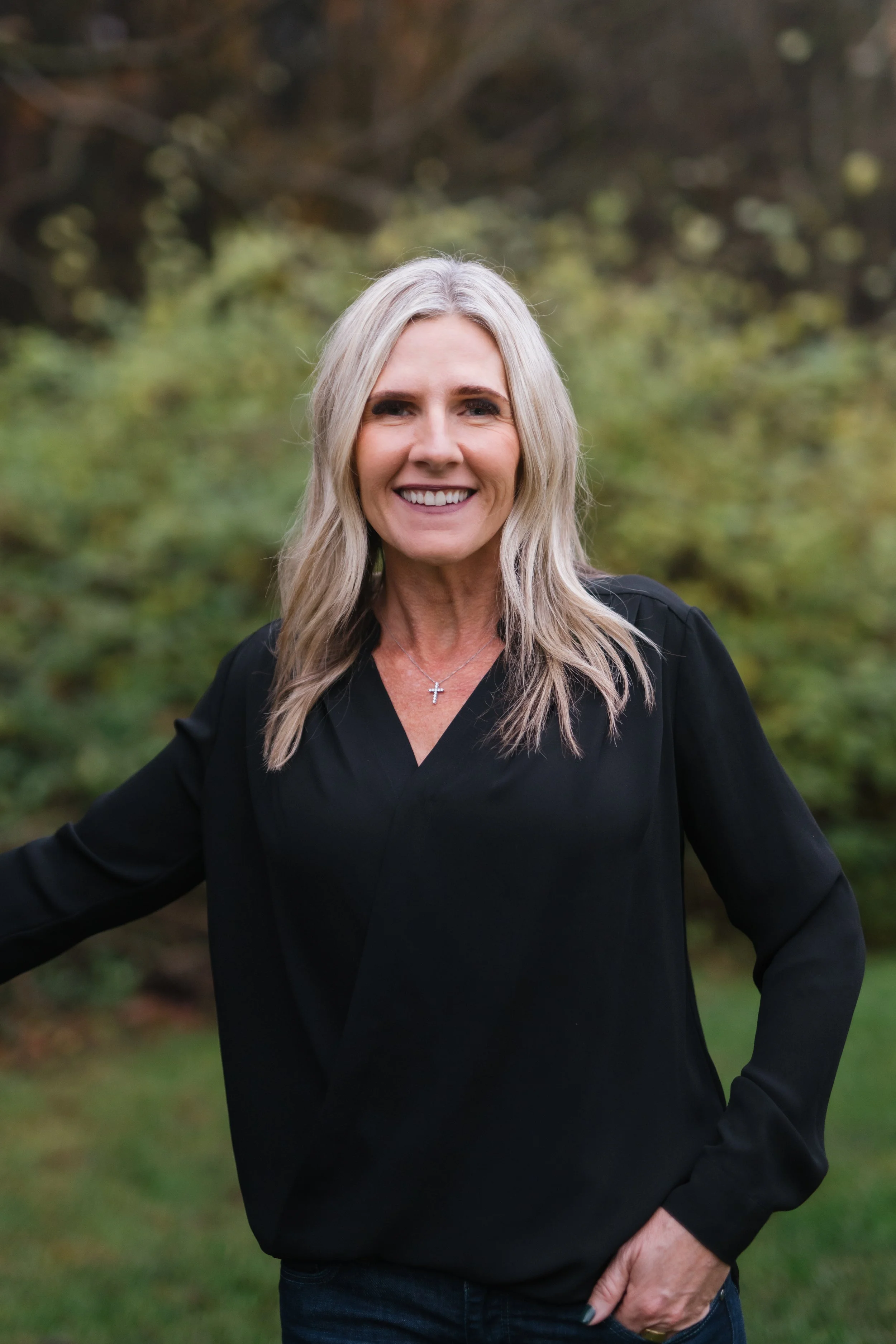 A woman with long, wavy gray hair smiling outdoors in a natural setting with trees and greenery in the background, wearing a black top and a cross necklace.