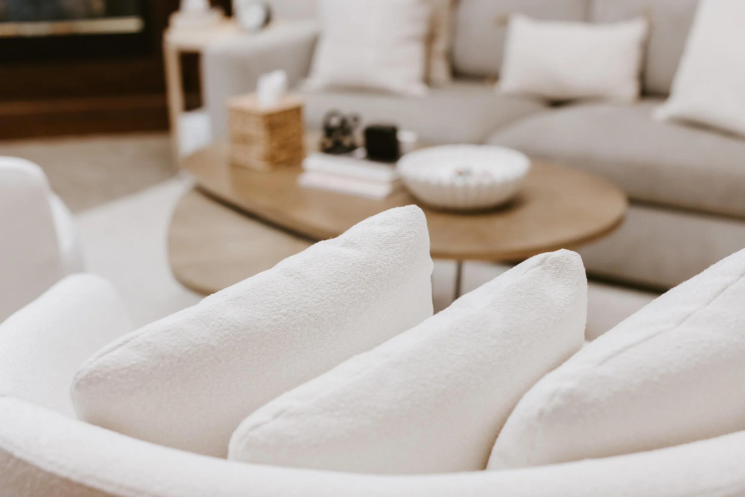 Close-up of white cushions on a sofa in a cozy living room setting with a round wooden coffee table in the background.