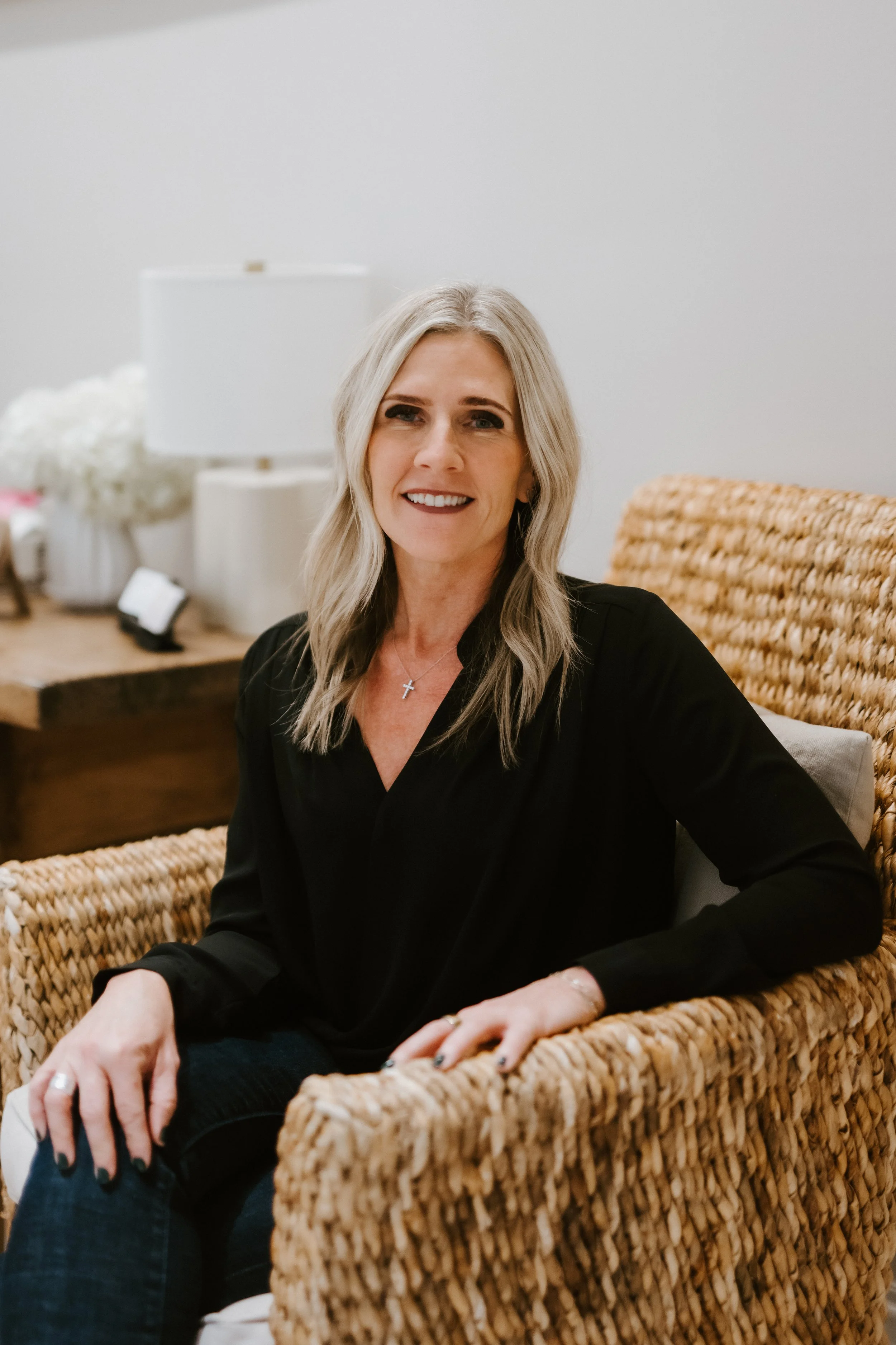 A smiling middle-aged woman with shoulder-length blonde hair, wearing a black top, sitting in a rattan chair in a cozy indoor setting.