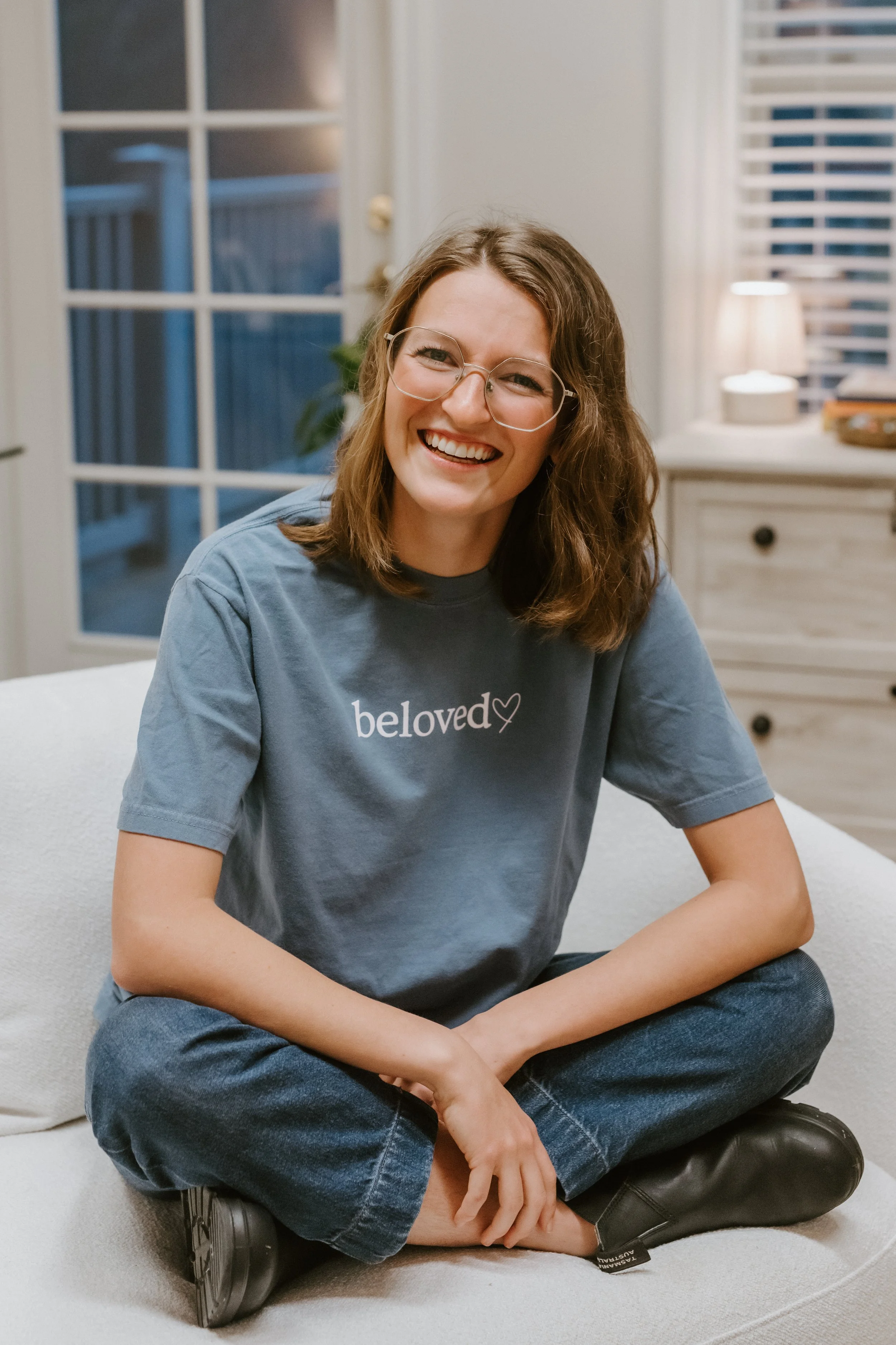 A smiling woman with glasses and shoulder-length brown hair sitting cross-legged on a white couch in a cozy living room, wearing a blue t-shirt with the word 'beloved' and a heart on it.