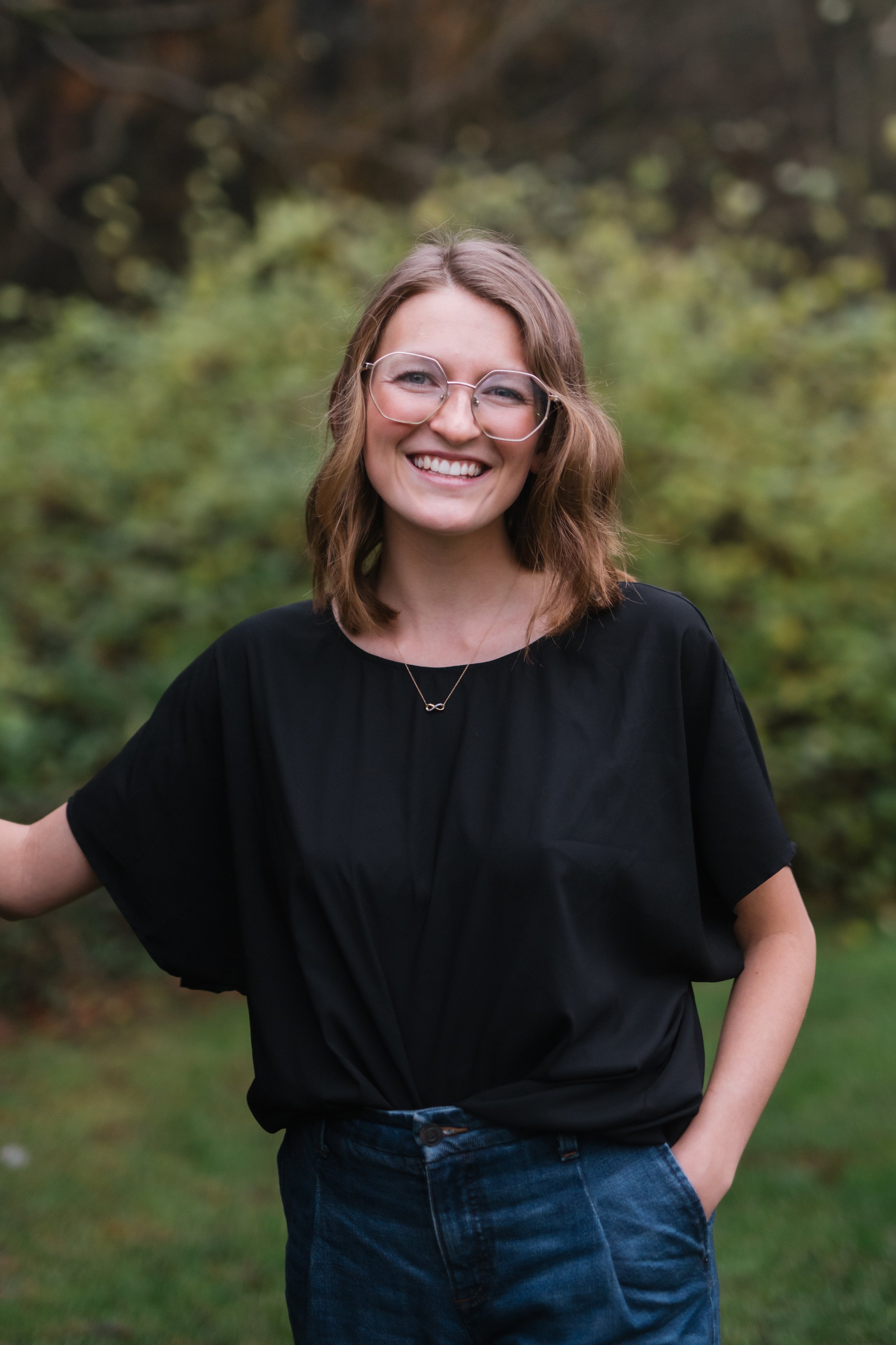 A smiling woman with shoulder-length light brown hair wearing glasses, a black t-shirt, and jeans outdoors in a green, wooded area.