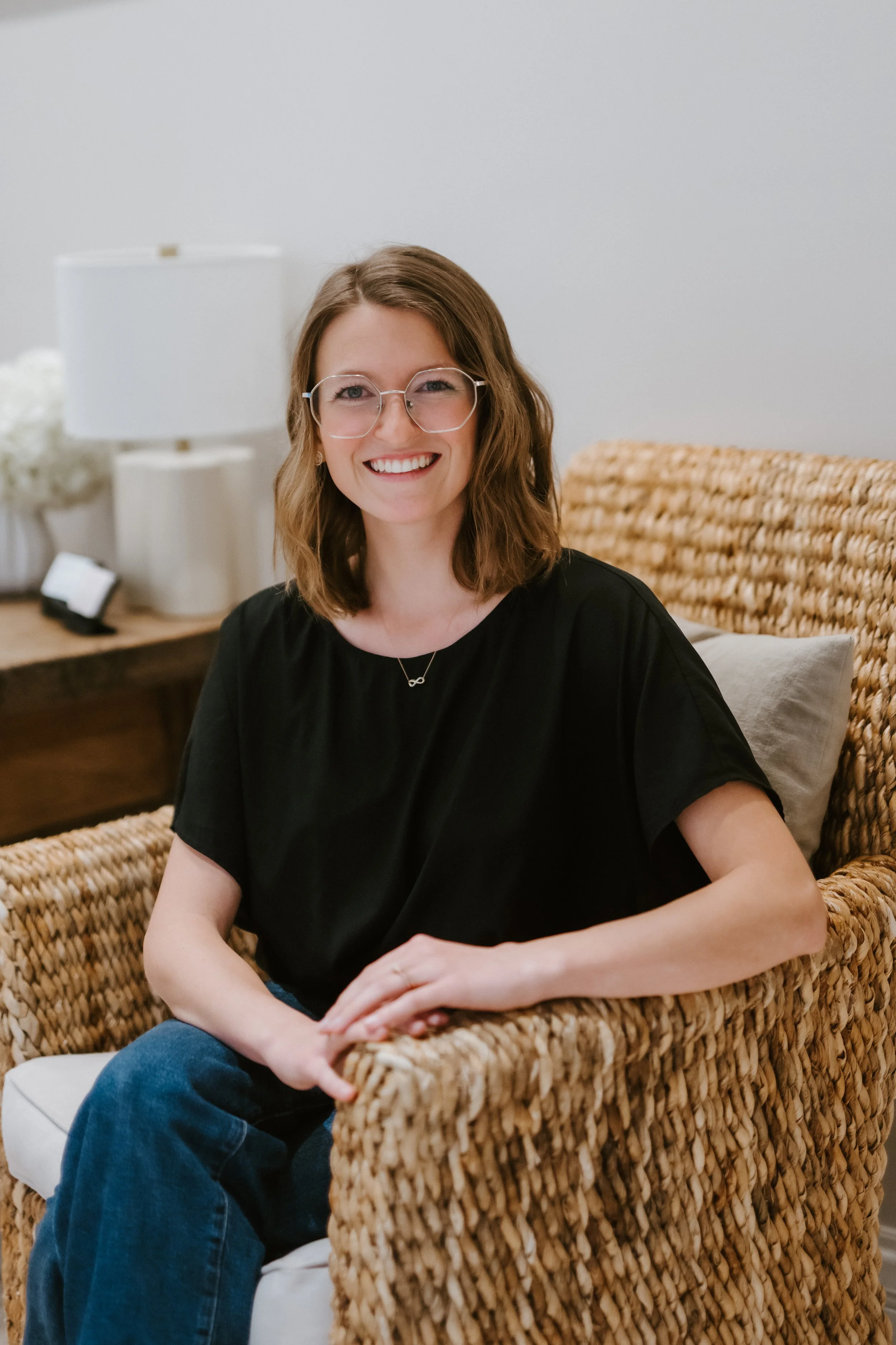 A woman with shoulder-length brown hair, glasses, and a black shirt, sitting on a wicker chair in a room with a wooden table, lamp, and books.