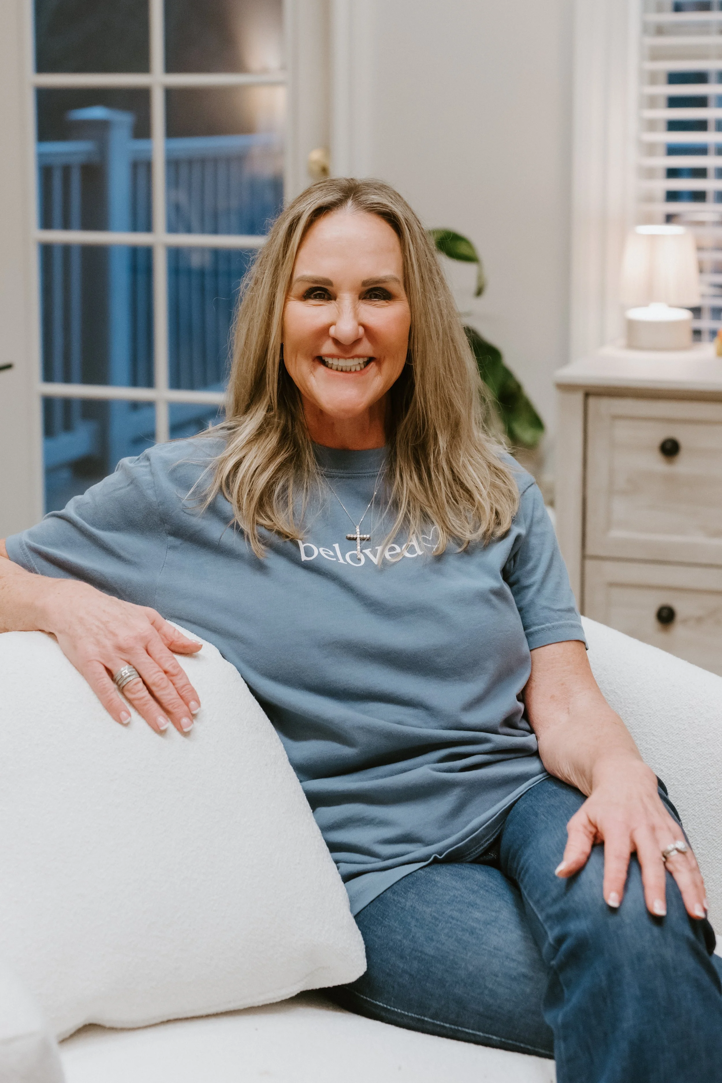 Smiling woman with long blonde hair, wearing a blue t-shirt and jeans, sitting on a white couch in a cozy living room.