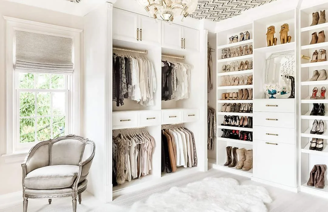 A walk-in closet with white shelves filled with shoes, boots, and handbags, a clothing rack with dresses and blouses, a window with a Roman shade, a plush armchair, and a white rug.