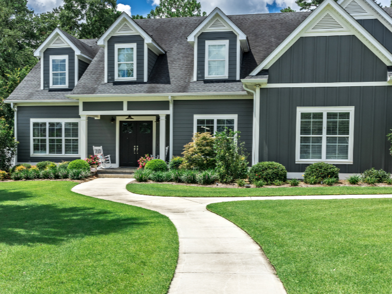 A modern two-story house with dark gray siding, white trim, and a front porch with a white bench. The house has multiple dormer windows and is surrounded by neatly landscaped green lawn and bushes under a partly cloudy sky.