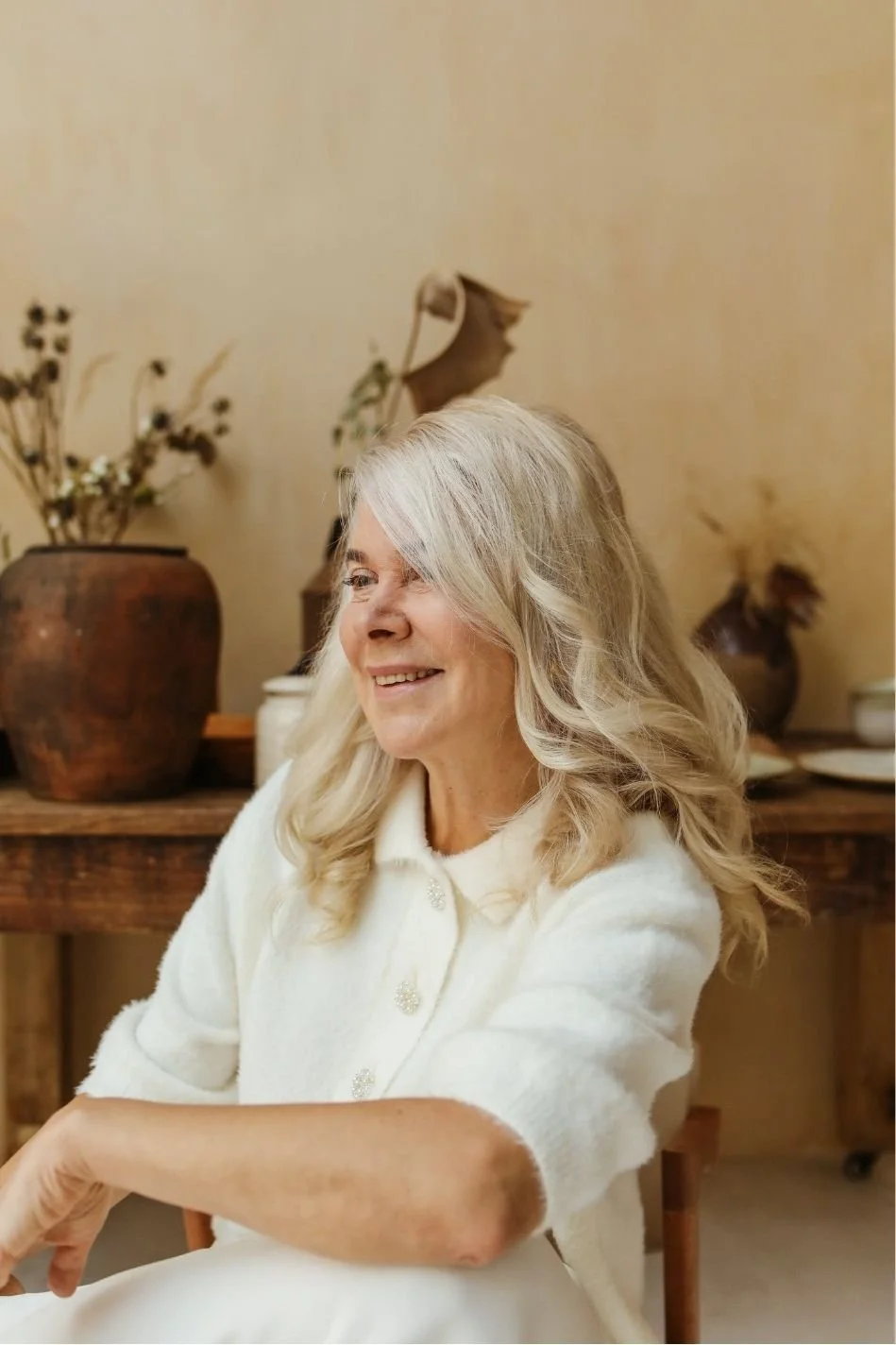 A smiling elderly woman with long gray hair sitting indoors, wearing a white sweater, with a wooden table and decorative vases with dried flowers in the background.