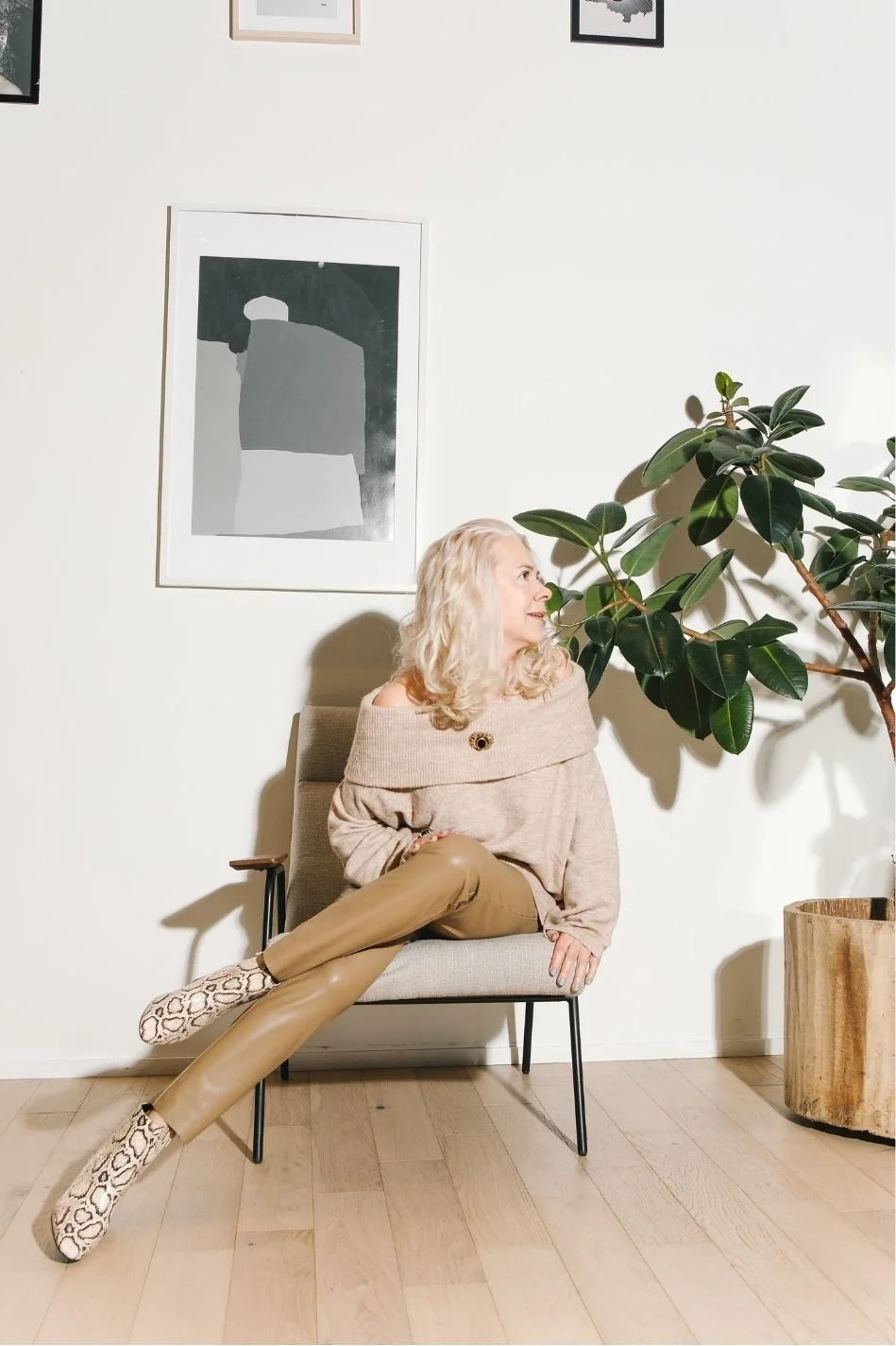A woman with blonde curly hair sitting on a beige chair with wooden armrest, wearing a beige off-the-shoulder sweater, tan pants, and patterned shoes, looking to her right near a large leafy green plant, with abstract artwork on the white wall behind her.