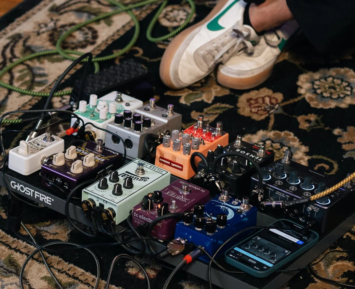 Guitar pedalboard with various effects pedals connected, placed on a patterned rug, with someone's legs and sneakers visible in the background.