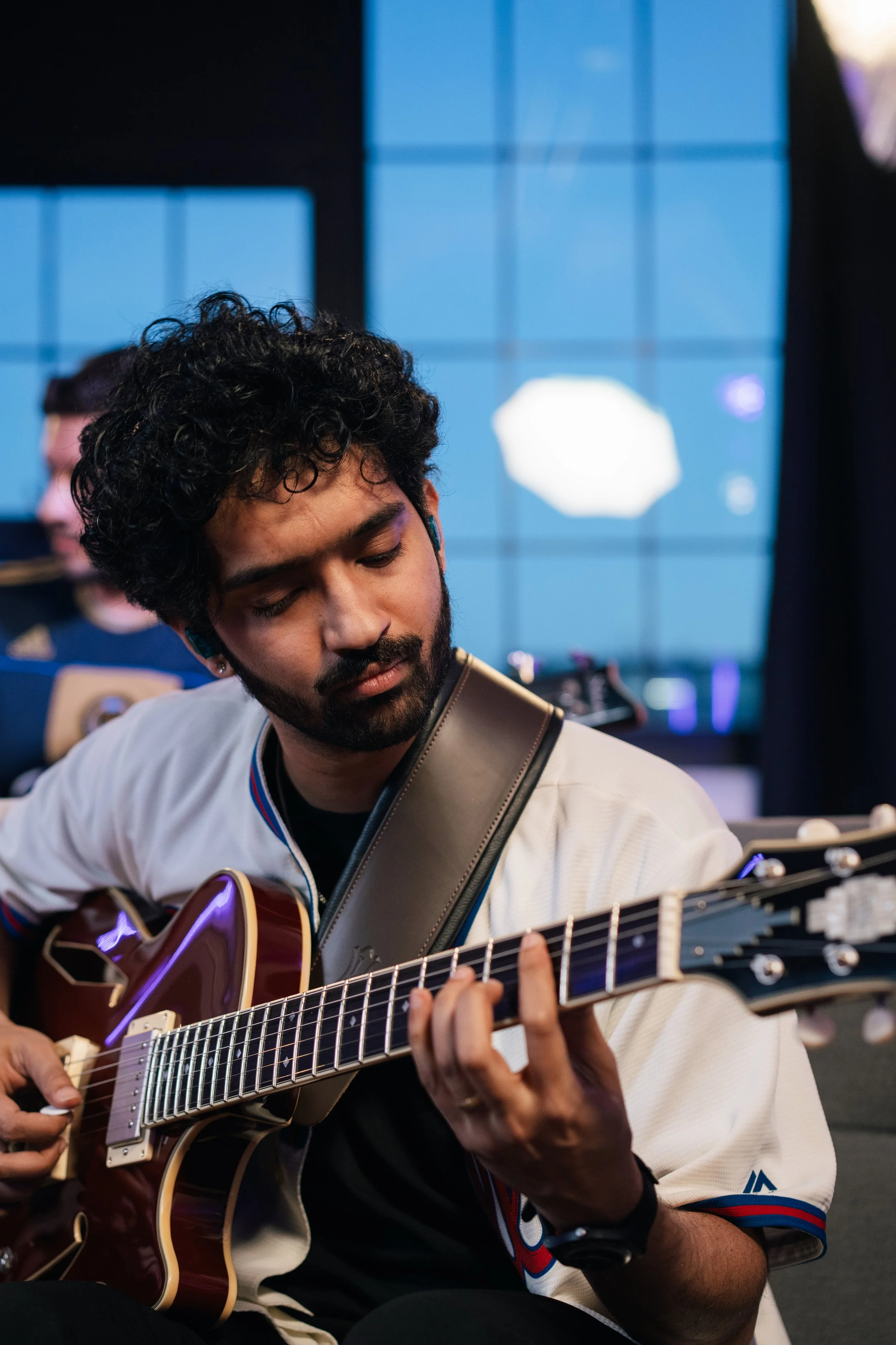 A young man with curly black hair and a beard playing an acoustic guitar while sitting in an indoor setting with large blue-lit screens in the background.