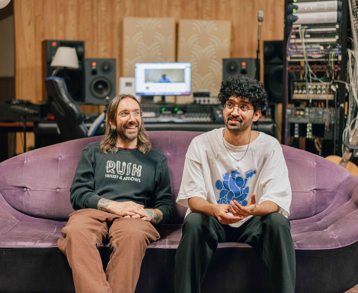 Two men sitting on a purple velvet couch in a music studio with audio equipment and mixing console in the background. They are smiling and engaged in conversation.