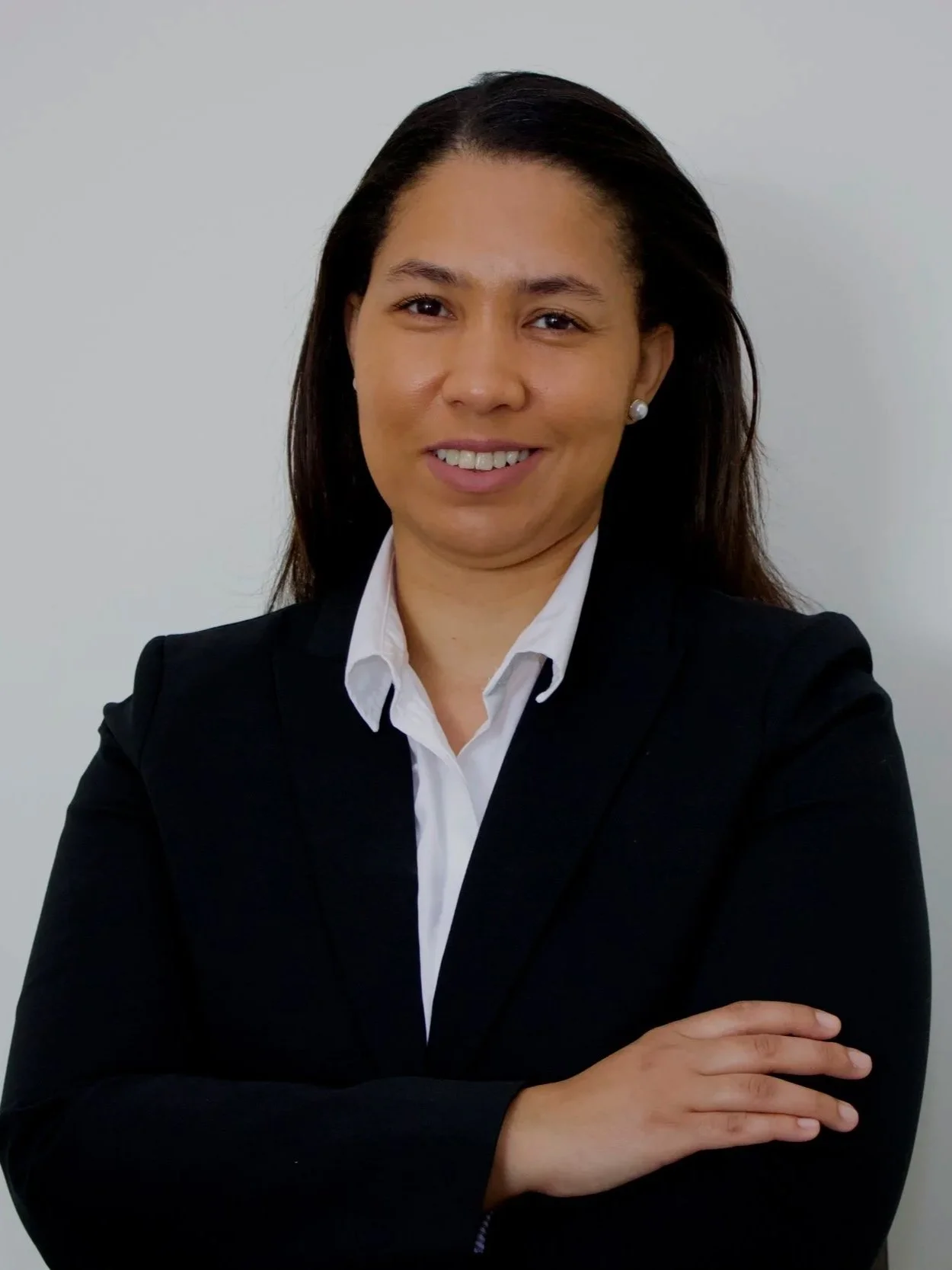 A professional woman with dark hair in a black blazer and white shirt, smiling and crossing her arms, standing against a light gray background.