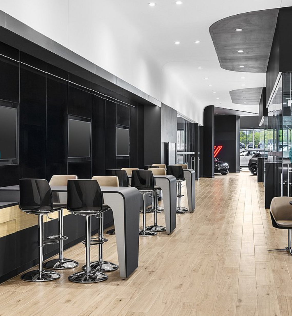 Empty modern car dealership waiting area with black and beige chairs, wooden flooring, large windows, and a few cars visible outside.