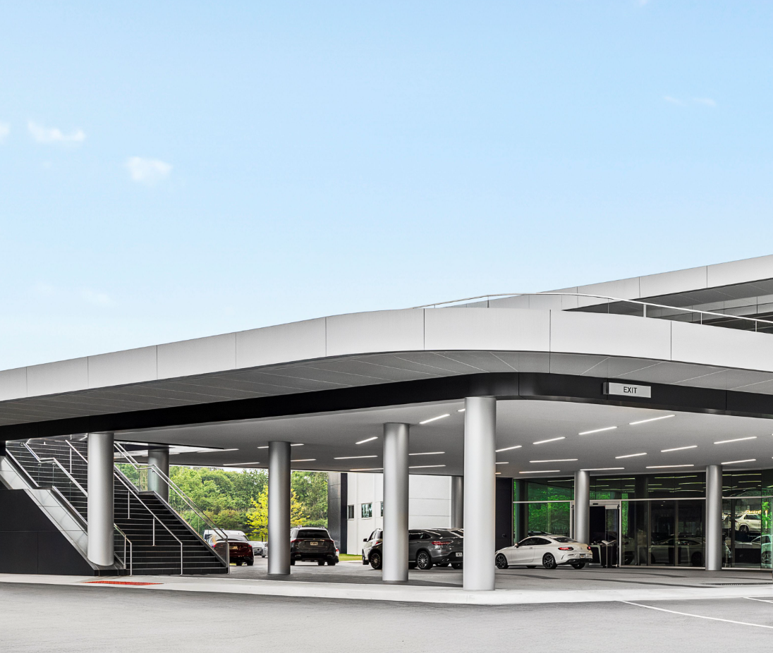 Car dealership with multiple cars parked under a modern, covered structure with a staircase on the left side and clear glass windows.