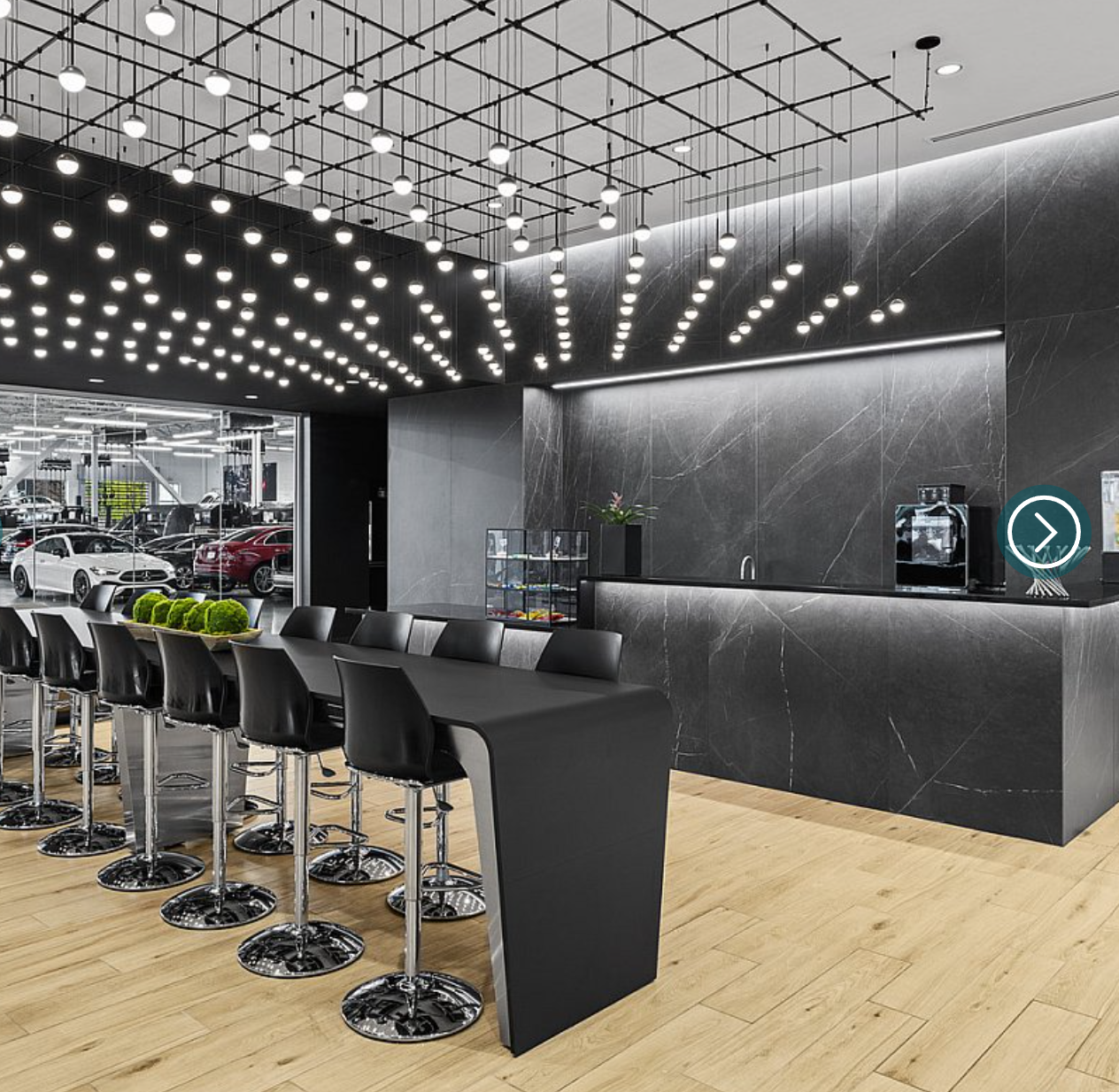 Modern car showroom interior with a long black counter, high black chairs, and ceiling lights, with a view of cars outside through large windows.
