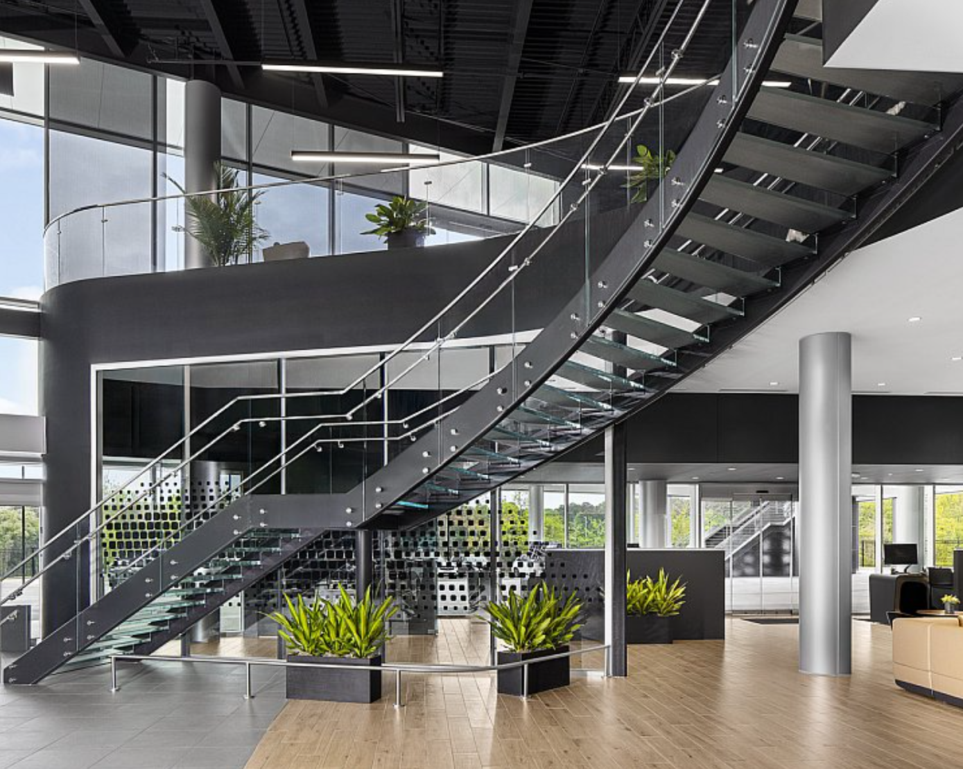 Modern office lobby with a staircase, glass railings, large windows, and indoor plants.