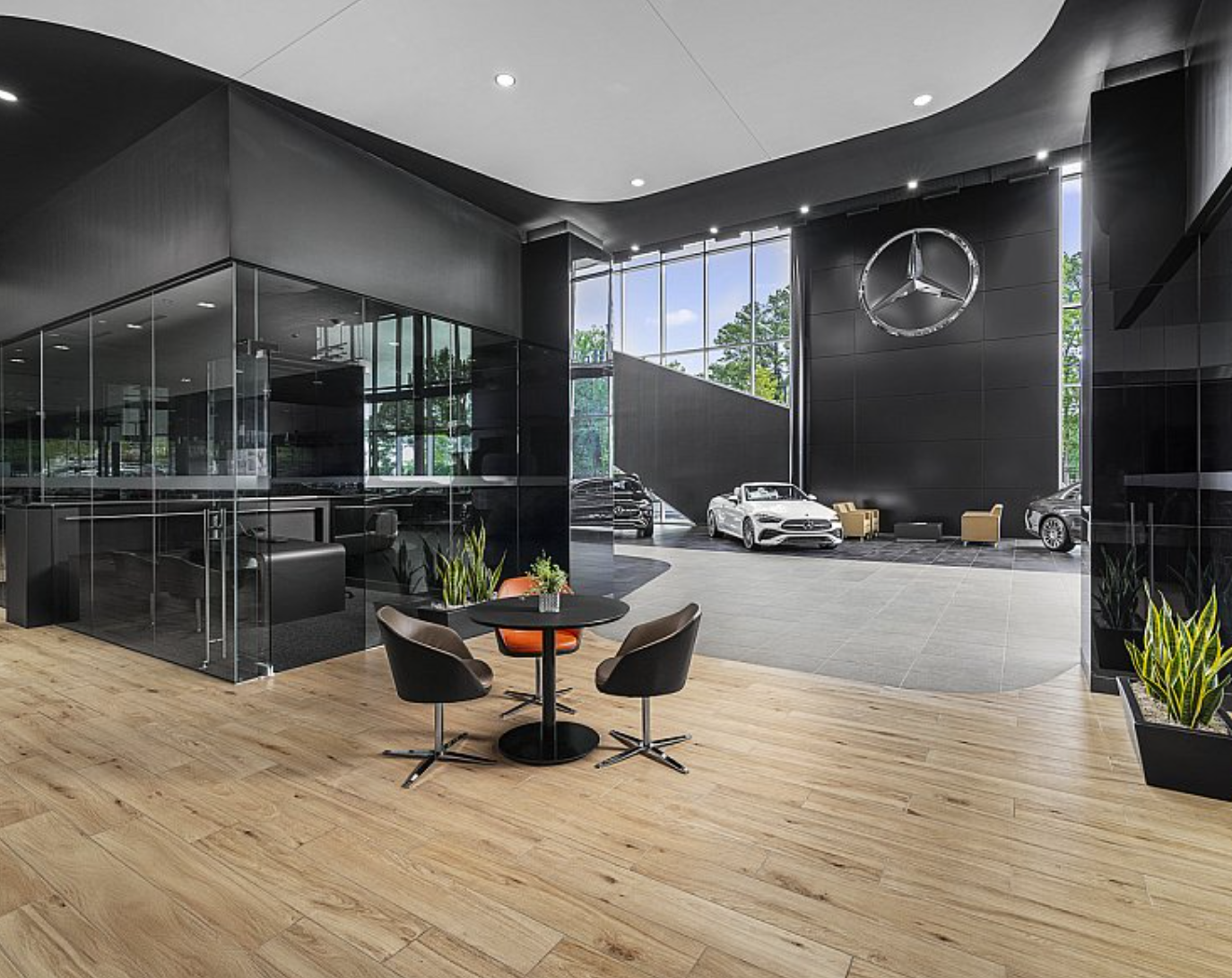 Modern Mercedes-Benz dealership showroom with cars displayed, glass walls, black walls, Mercedes logo, seating area, and wood flooring.
