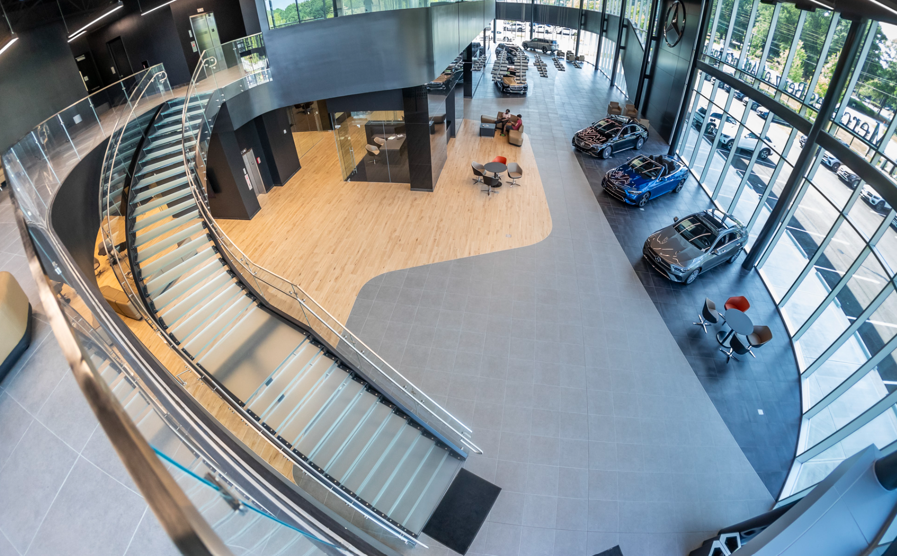 An interior view of a modern car dealership showroom with several cars on display near floor-to-ceiling windows. There is a curved staircase with glass railing leading to an upper level and a seating area with tables and chairs.