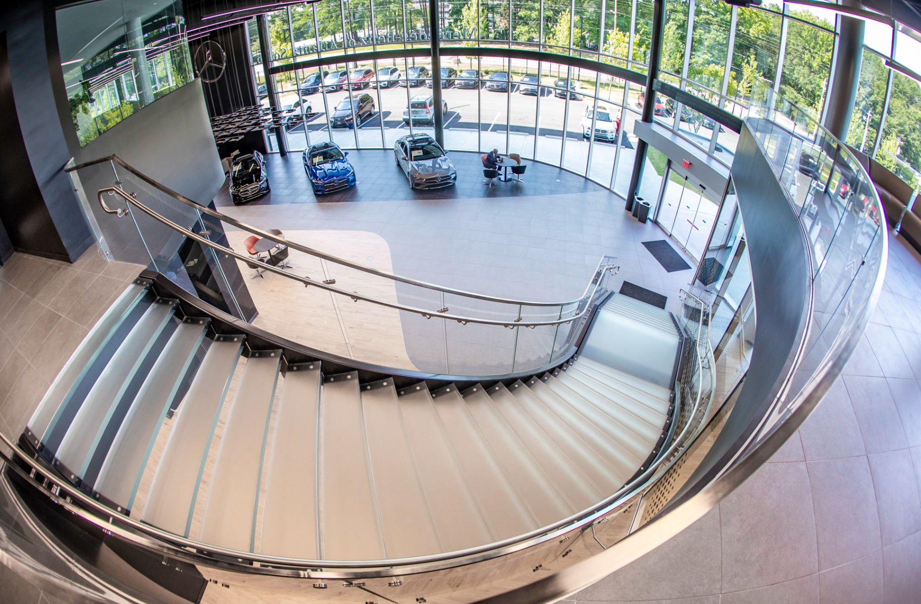 View from the upper floor of a car dealership showroom, showing the staircase leading down to the main level, where three cars are displayed near large glass windows. Outside, there are parked cars and greenery.